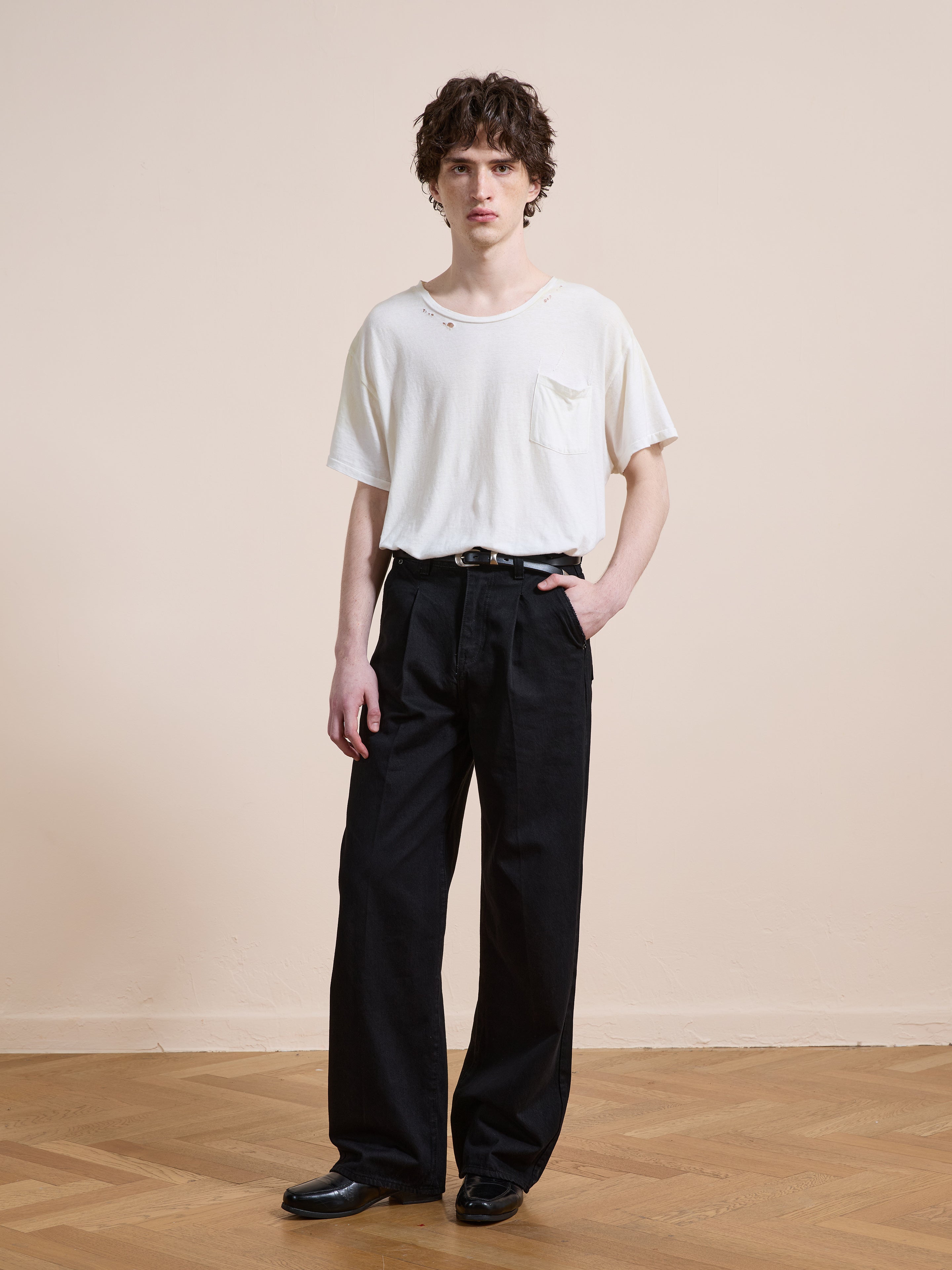 Indoors on a wooden floor, a person wears Found Pleated Relaxed Fit Jeans with a black belt, paired with a white t-shirt and black shoes, one hand in their pocket.