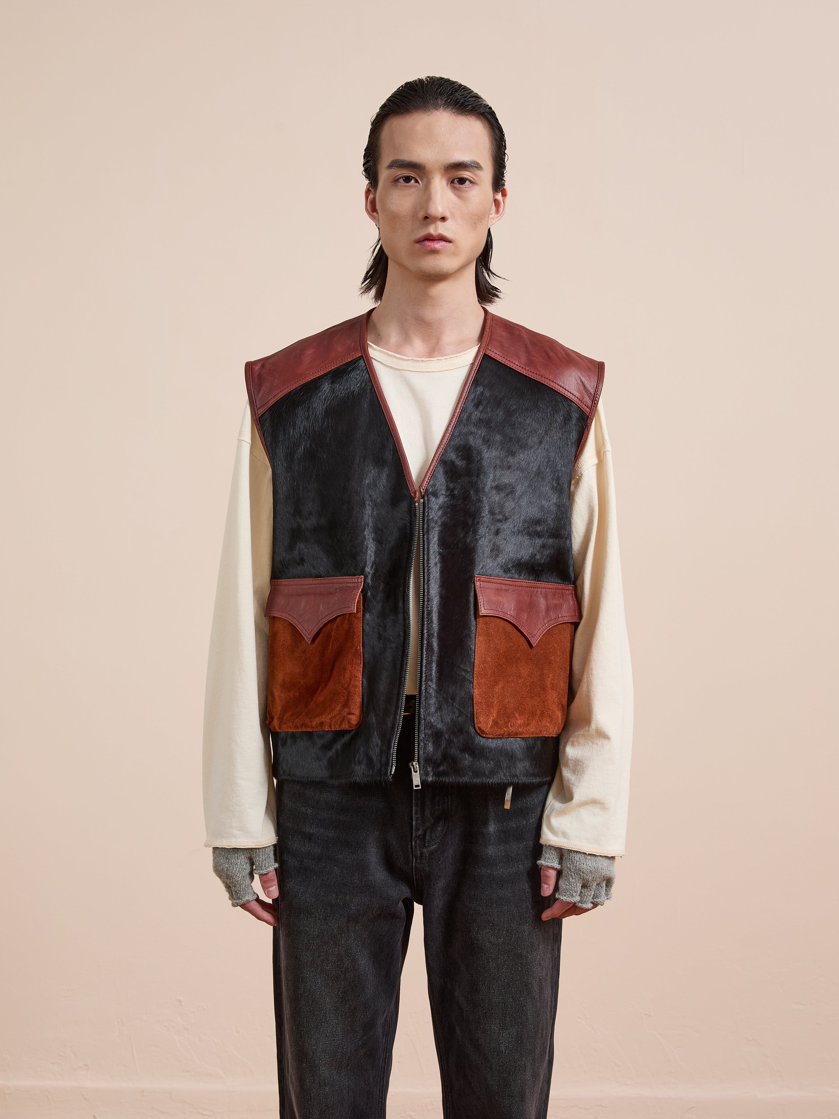 A person stands facing forward, wearing Found’s Fur Western Leather Vest with large pockets over a cream long-sleeve shirt, plus fingerless gloves and dark jeans, against a plain beige background.