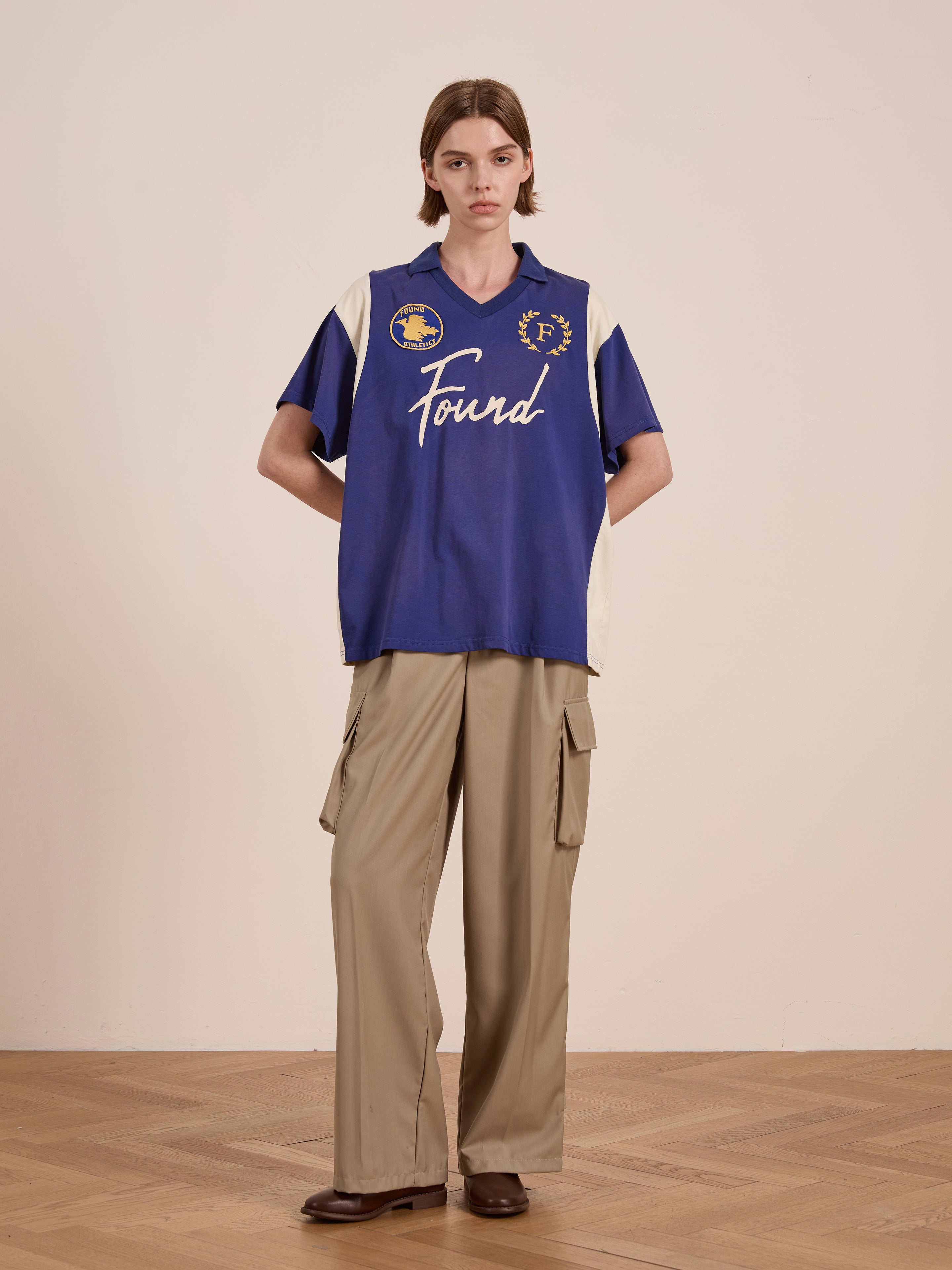 Indoors, a person wears a Premier League Jersey by FOUND, echoing vintage sportswear with blue and beige short sleeves. Paired with beige cargo pants and brown shoes, the outfit exudes a relaxed yet classic sporty vibe.