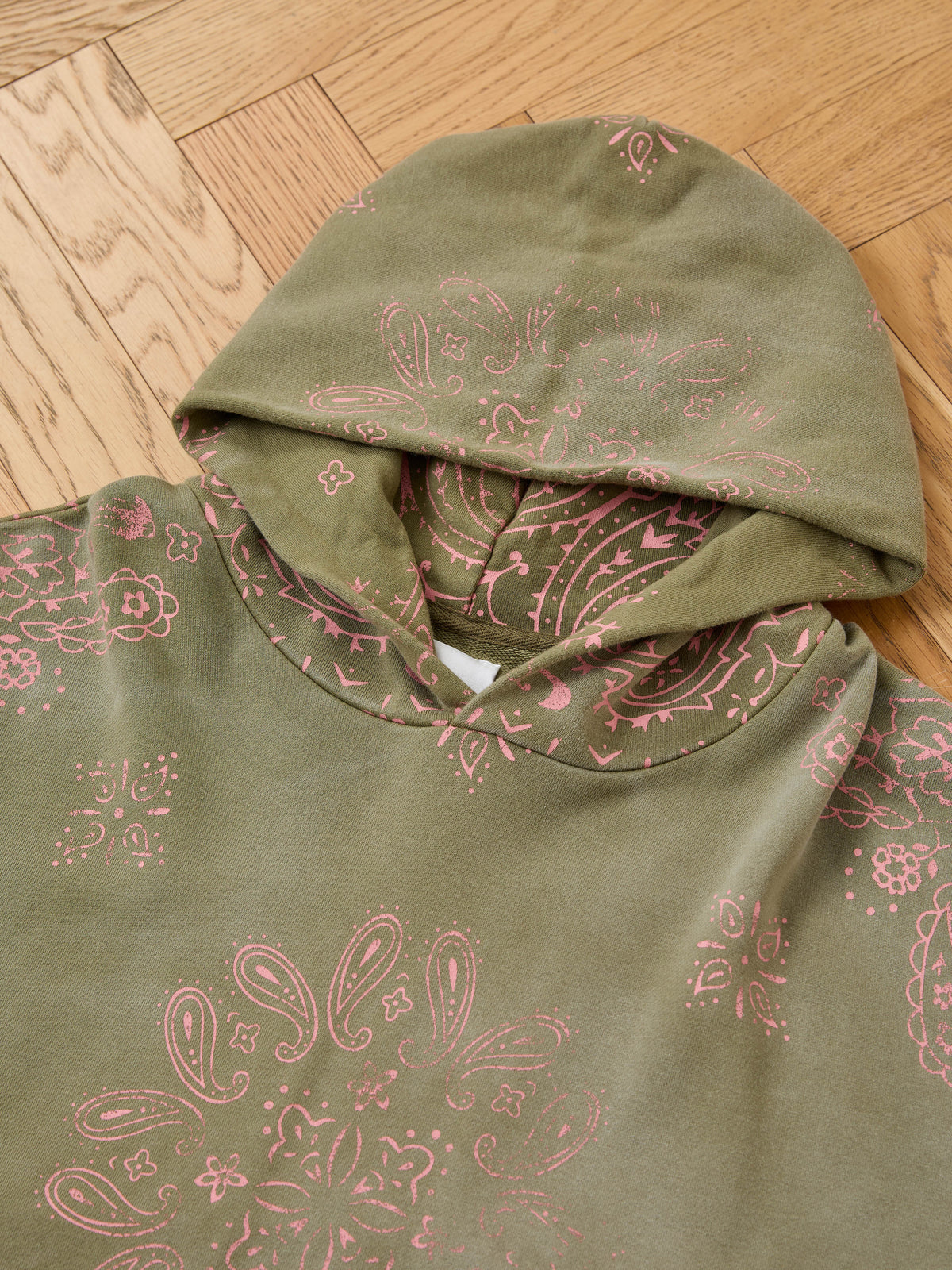 The FOUND Printed Paisley Hoodie in olive green with a pink paisley design is a unisex pullover shown laid flat on a wooden floor.
