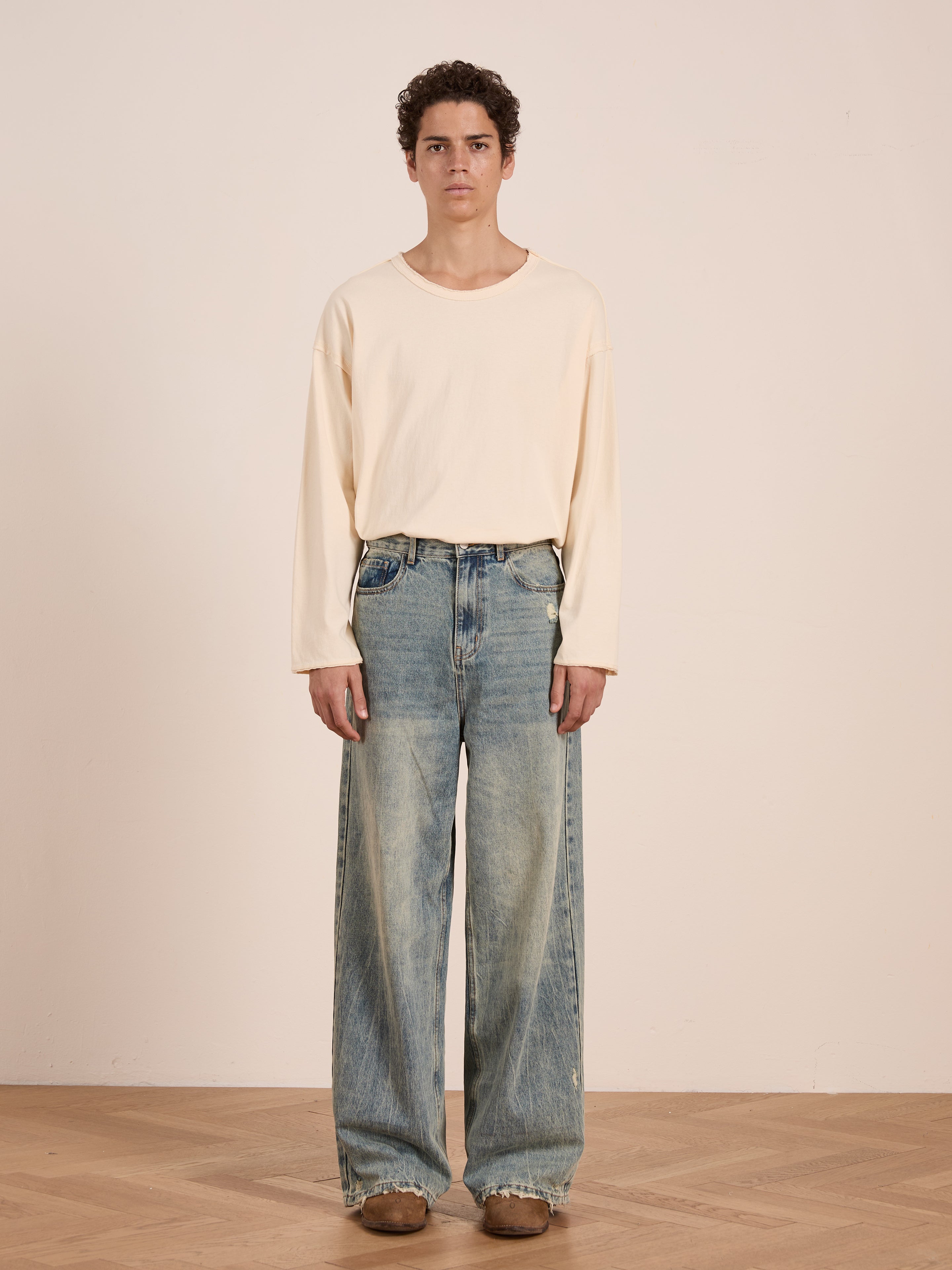 Indoors against a plain wall, a person wears FOUND’s Reversed LS Tee—a loose unisex long sleeve with a distressed neckline—paired with wide-leg faded jeans and brown shoes.