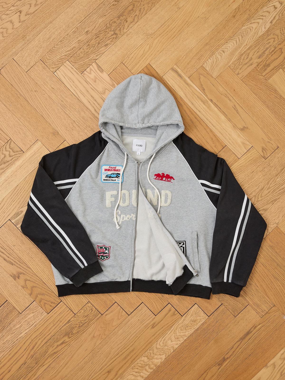 Gray and black Racer Hoodie by FOUND features embroidered patches, white sleeve stripes, and “FOUND sports” text on the front, shown laid flat on a wooden floor.
