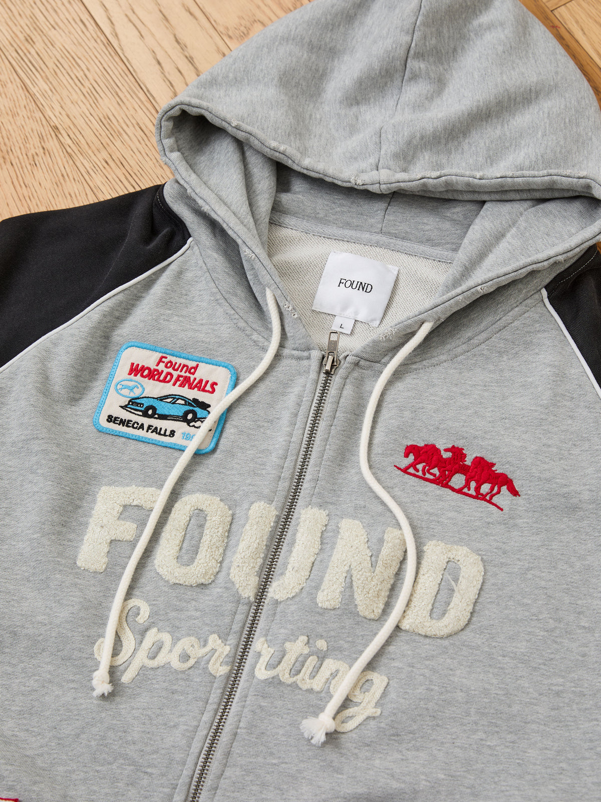 FOUND Racer Hoodie in gray with “FOUND Sporting” text, white drawstrings, black sleeves, red horse logo, and blue “Found World Finals” chest patch on wood floor.