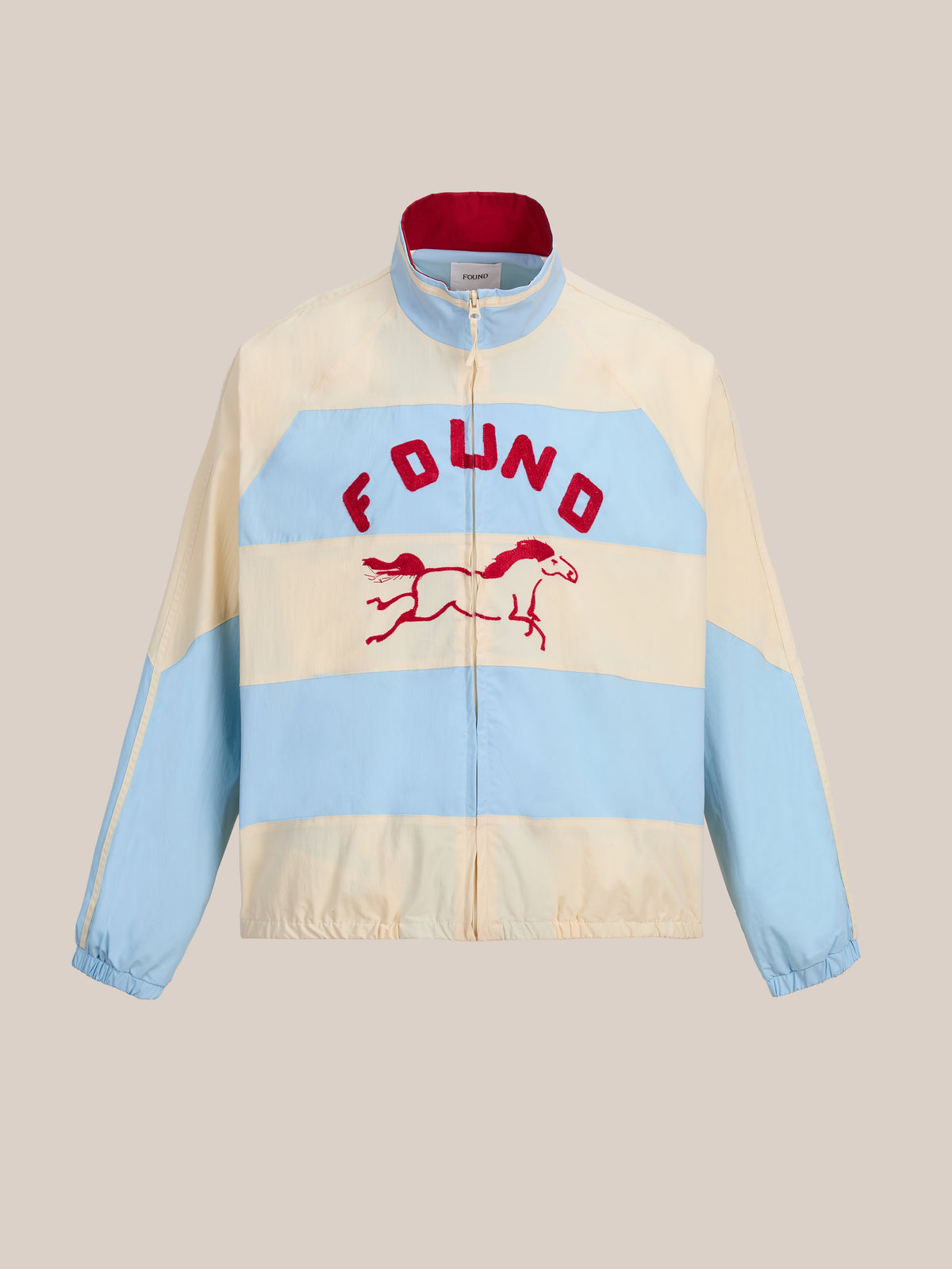 The FOUND Racing Striped Windbreaker features light blue and cream color-blocked stripes, bold red “FOUND” lettering, and a red embroidered horse graphic on the front—offering a modern, unisex twist on the classic vintage sportswear jacket for effortless everyday style.