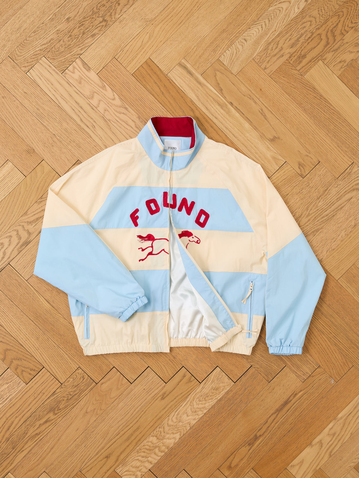 A FOUND Racing Striped Windbreaker in light blue and beige, featuring “FOUND” and two red running dogs printed on the front, is laid flat on a wooden floor. The unisex lightweight jacket offers a nod to vintage sportswear style.