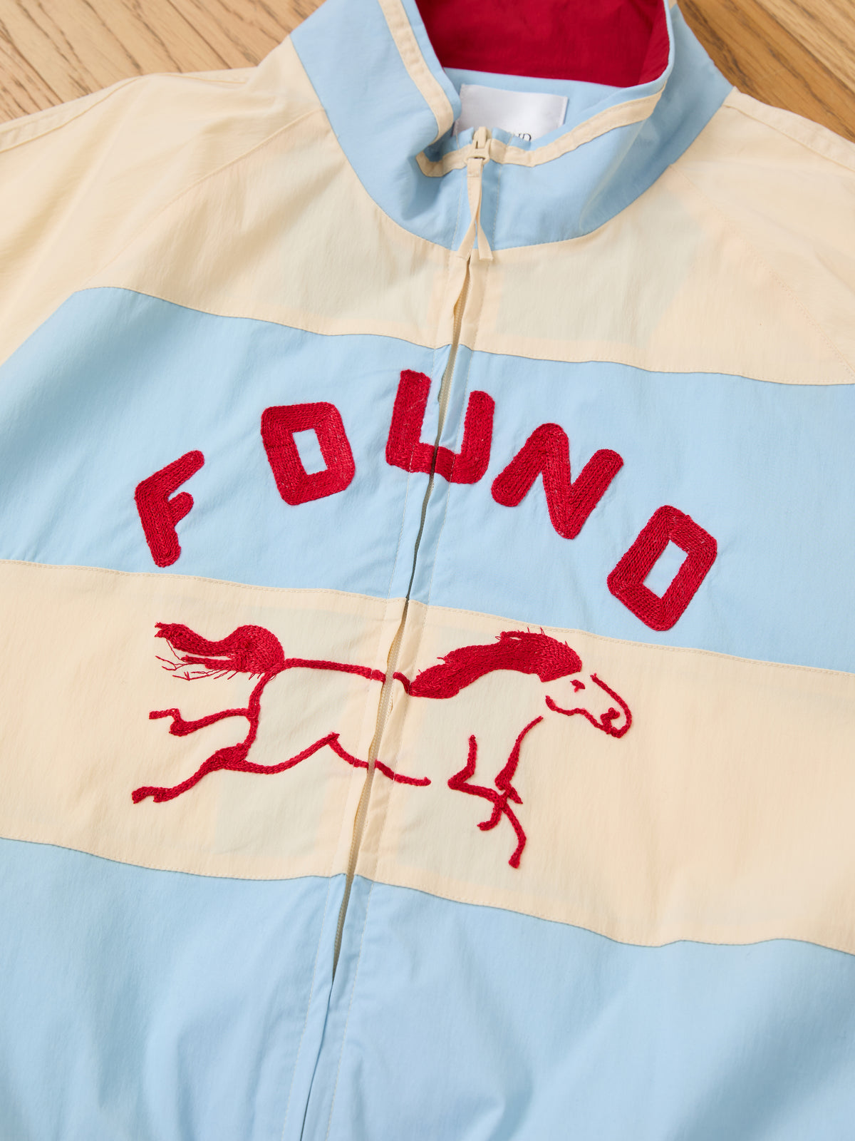 The FOUND Racing Striped Windbreaker, a unisex lightweight jacket inspired by vintage sportswear, features light blue and cream color-blocking with red embroidered “FOUND” lettering and a running horse on the front, shown displayed on a wooden surface.