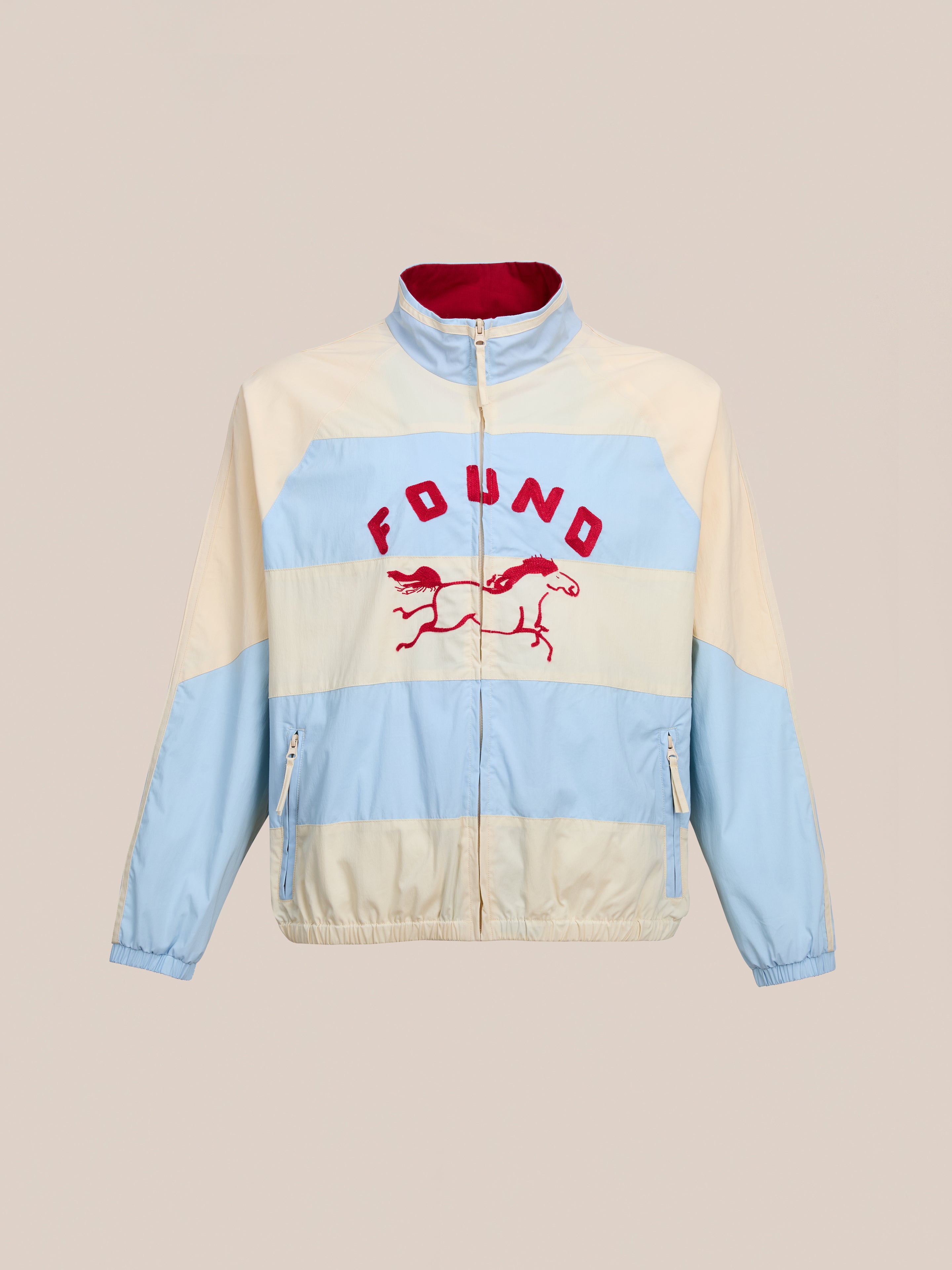 The FOUND Racing Striped Windbreaker is a vintage-inspired jacket with beige and light blue panels, red "FOUND" text and horse graphic on the chest, a front zipper, and elastic cuffs.