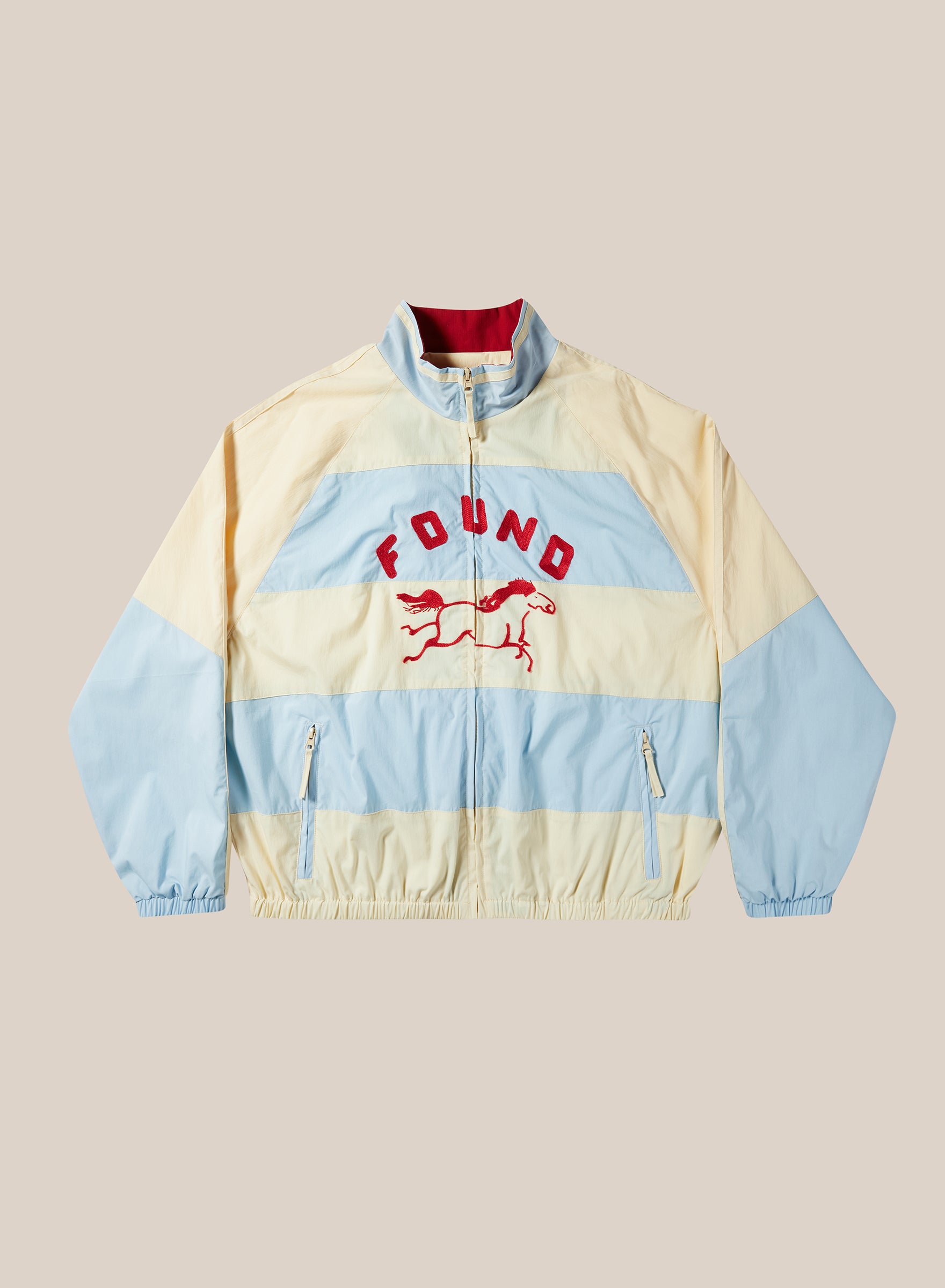 The Racing Striped Windbreaker by FOUND is a light blue and cream color-blocked jacket with a front zipper, "FOUND" text, and two running horse graphics on the chest, shown against a plain background.