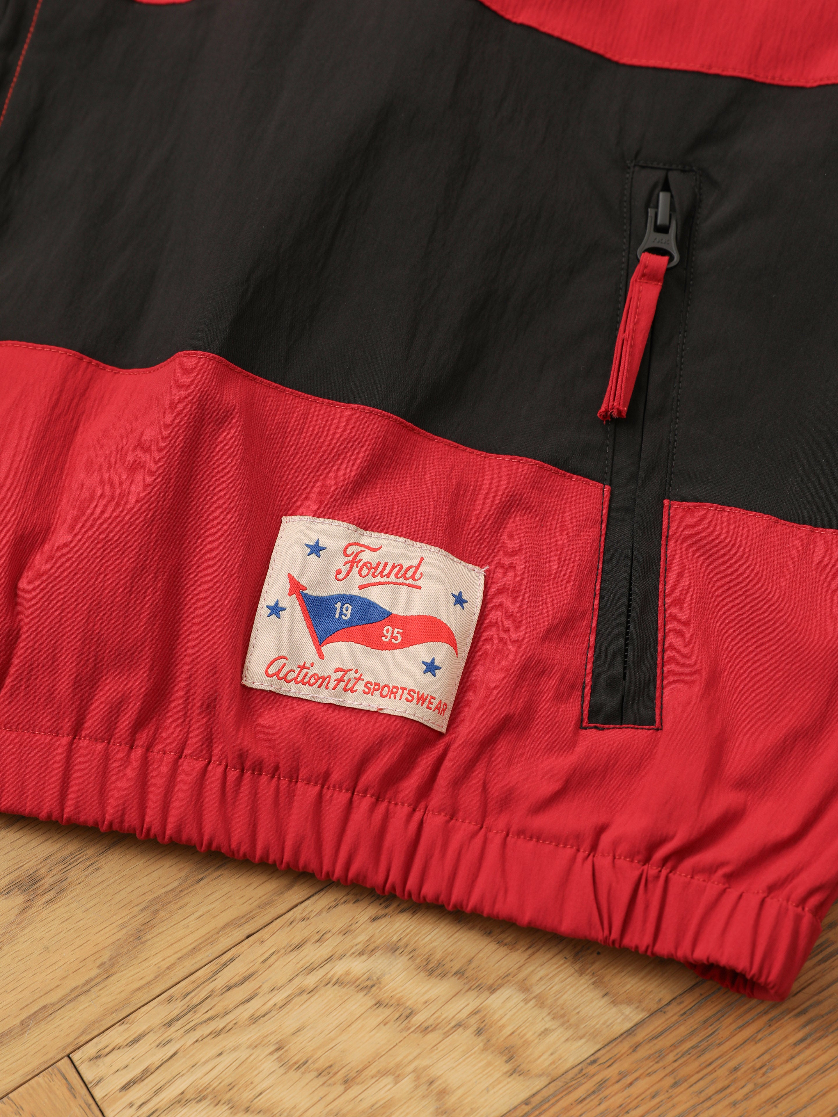 Close-up of a FOUND Racing Striped Windbreaker in red and black with a zipper pocket and "Found 1995 ActionFit Sportswear" label, displayed on a wooden floor.