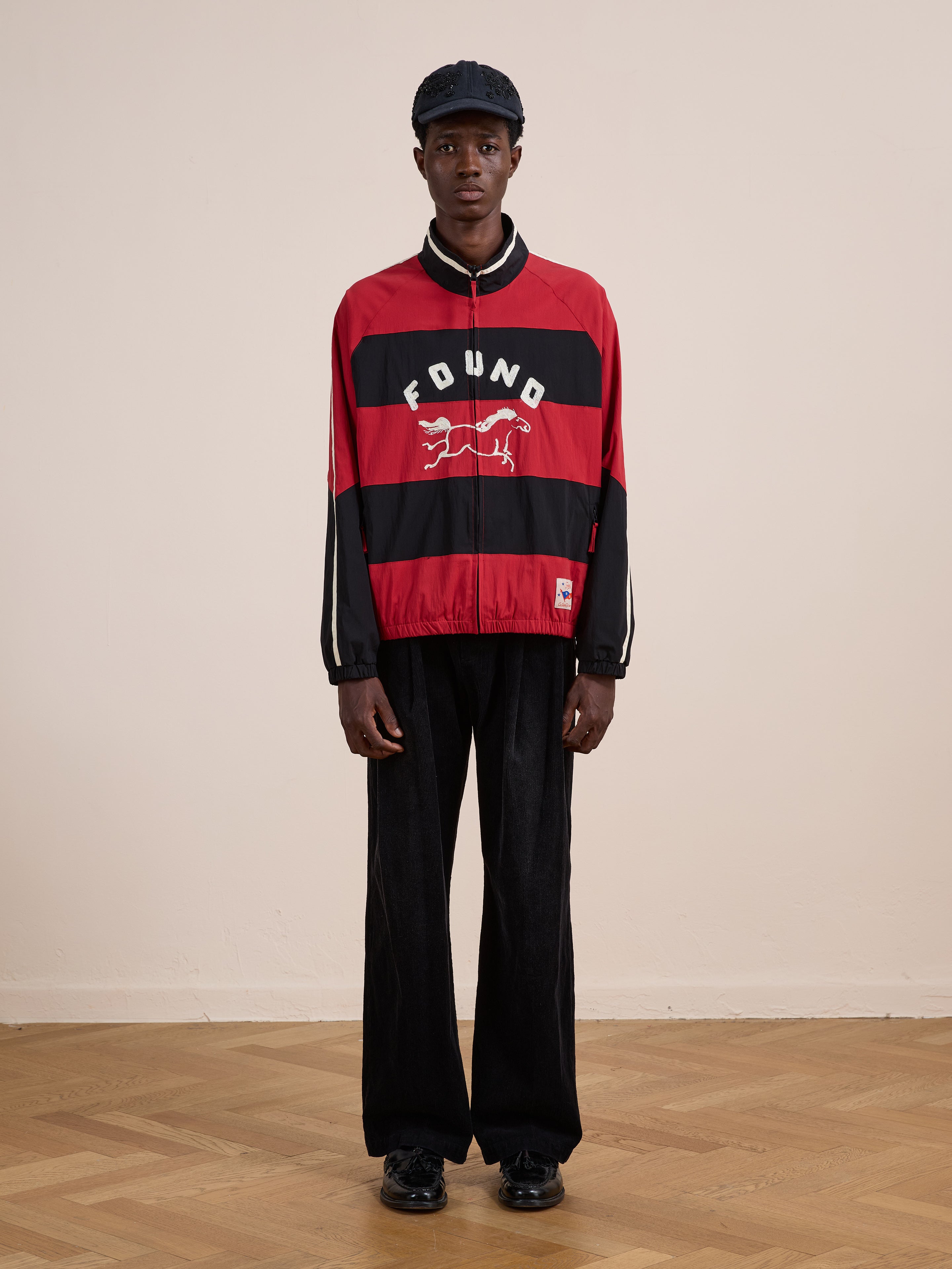 Indoors, a person wears the FOUND Racing Striped Windbreaker featuring a front graphic, paired with black pants, shoes, and cap against a plain background.