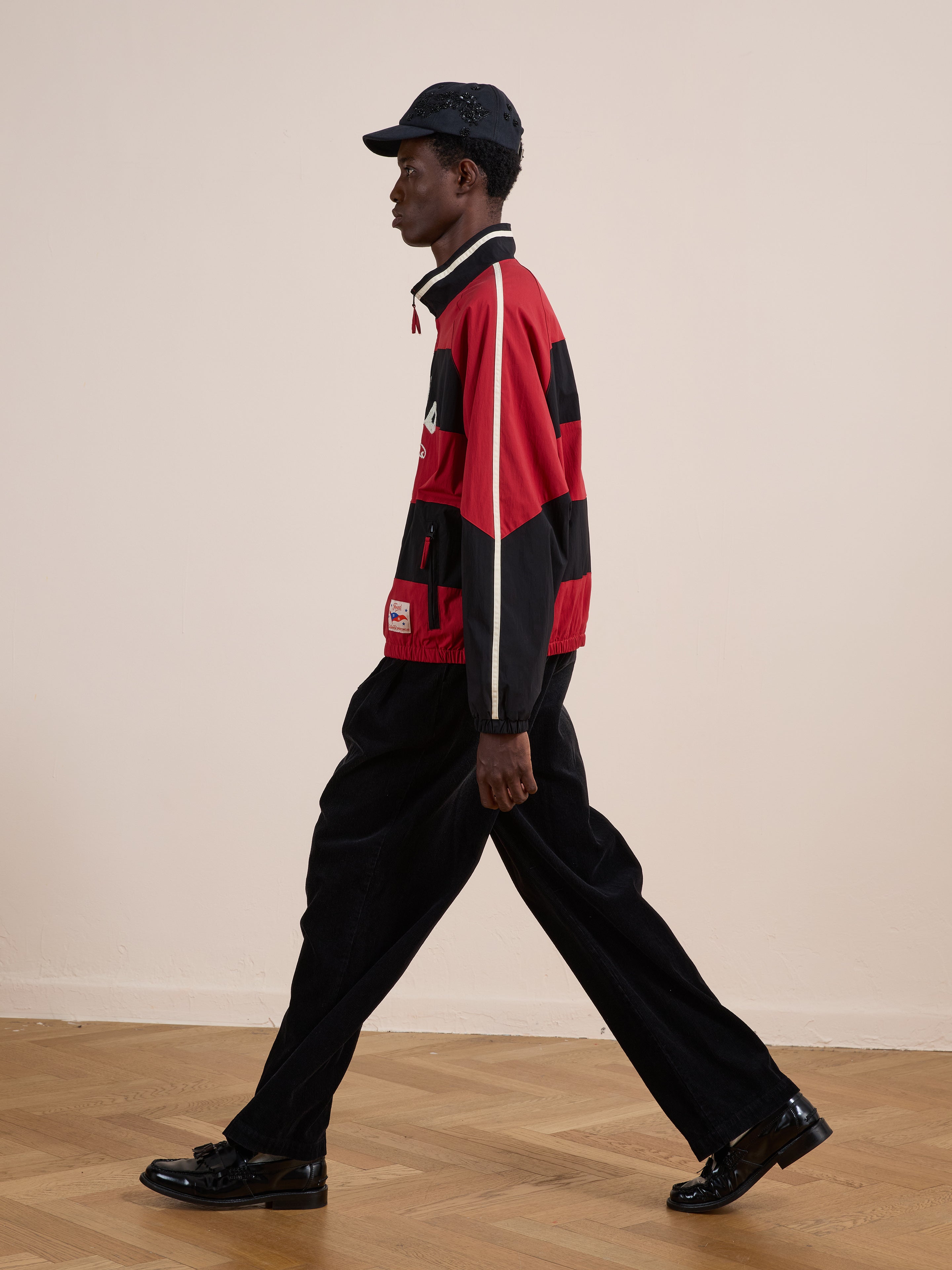 A man in a FOUND Racing Striped Windbreaker, black wide-leg pants, and a black cap walks indoors on a wooden floor.