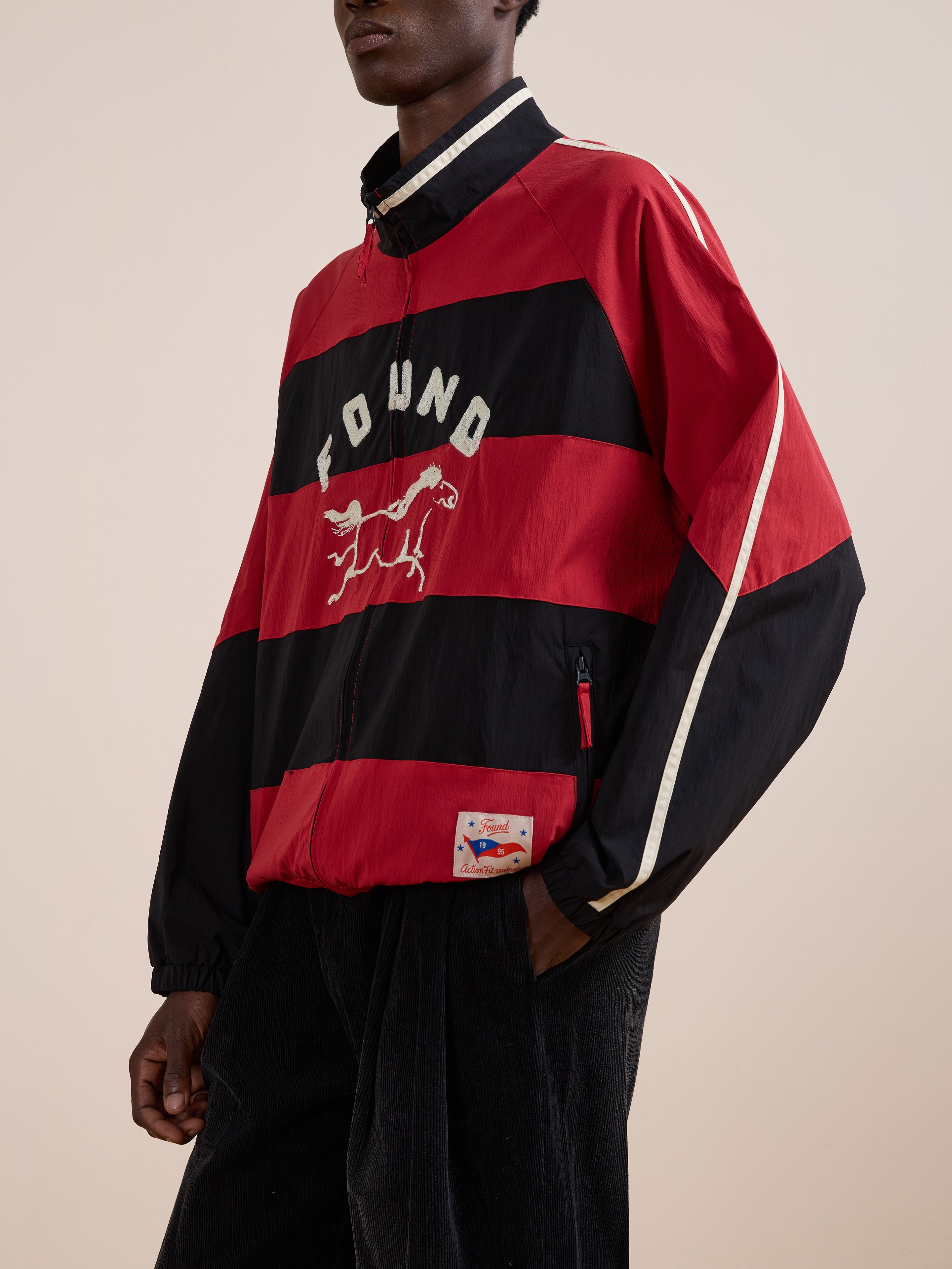 A person wears the FOUND Racing Striped Windbreaker, a vintage sportswear-inspired red and black jacket featuring "FOUND" and a horse graphic on the chest, standing against a plain background.