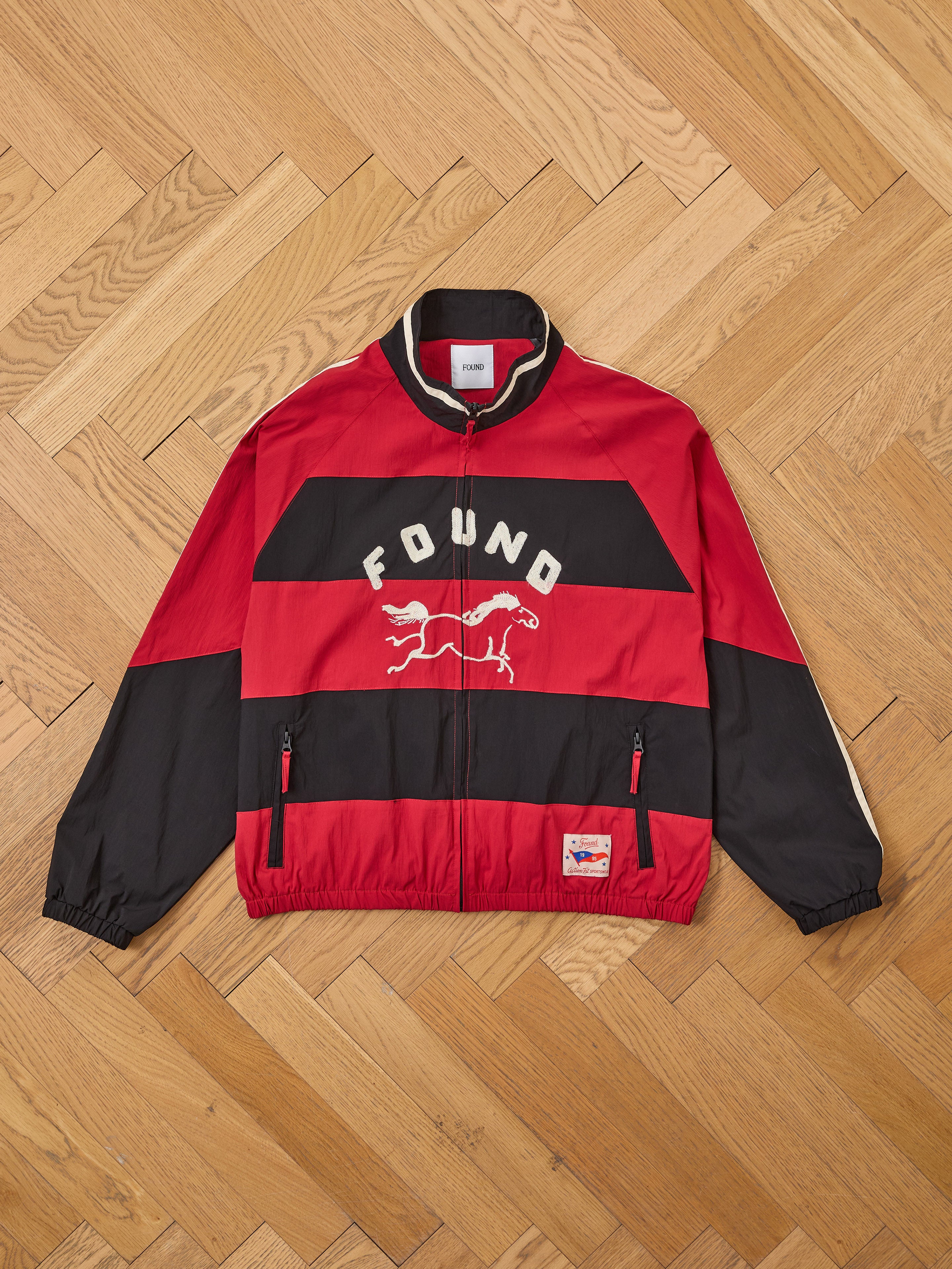 The FOUND Racing Striped Windbreaker is a unisex red and black jacket with "FOUND" and a running animal graphic on the chest, vintage sportswear-inspired, shown on a wooden parquet floor.