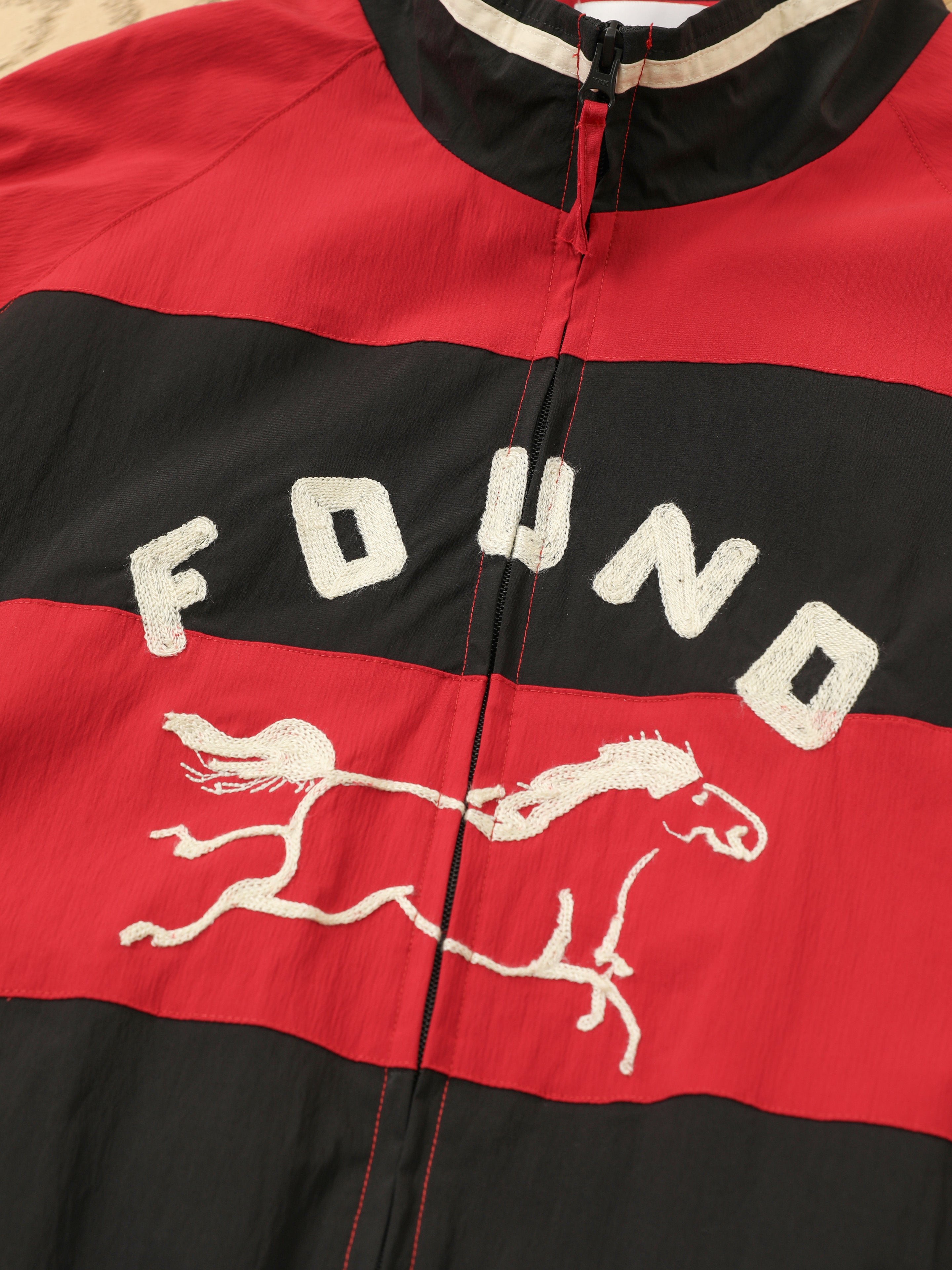 The FOUND Racing Striped Windbreaker is a red and black unisex jacket with "FOUND" and an embroidered running horse on the front, blending vintage sportswear appeal with a timeless athletic style.