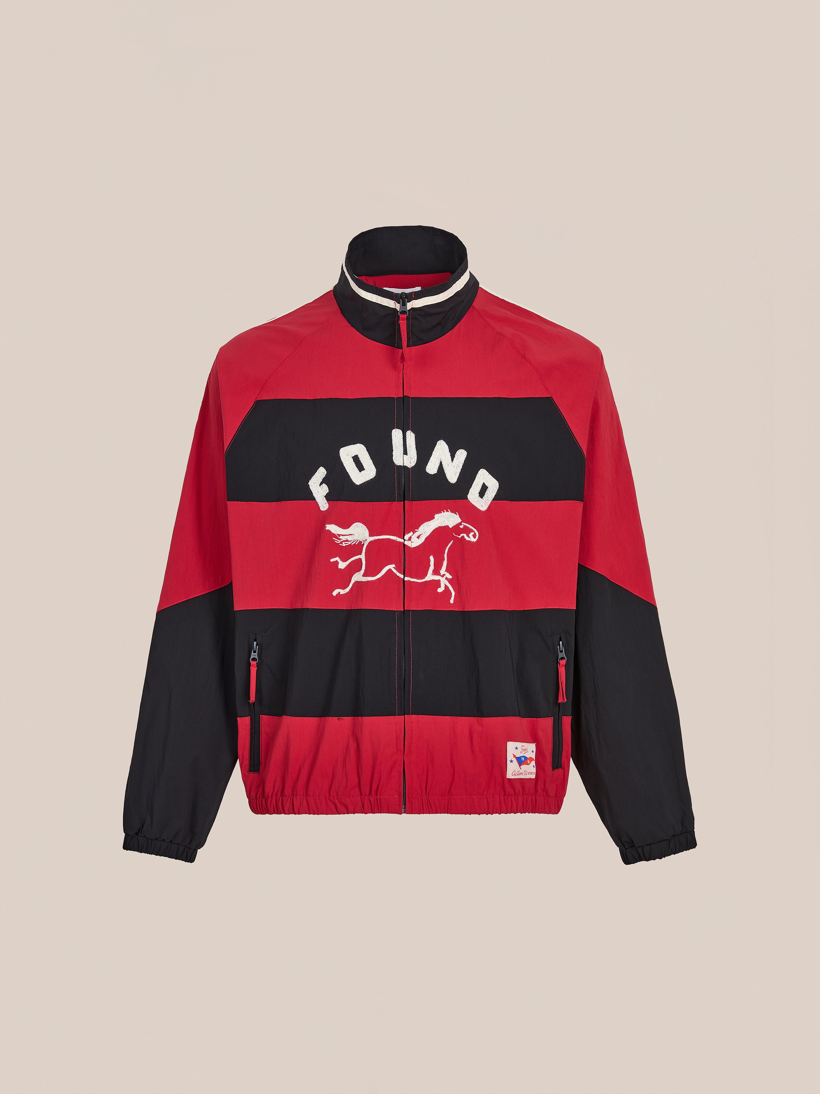 The FOUND Racing Striped Windbreaker is a red and black jacket with racing stripes, a horse graphic and "FOUND" on the front, two zippered pockets, and a small hem patch, inspired by vintage sportswear for a classic unisex look.