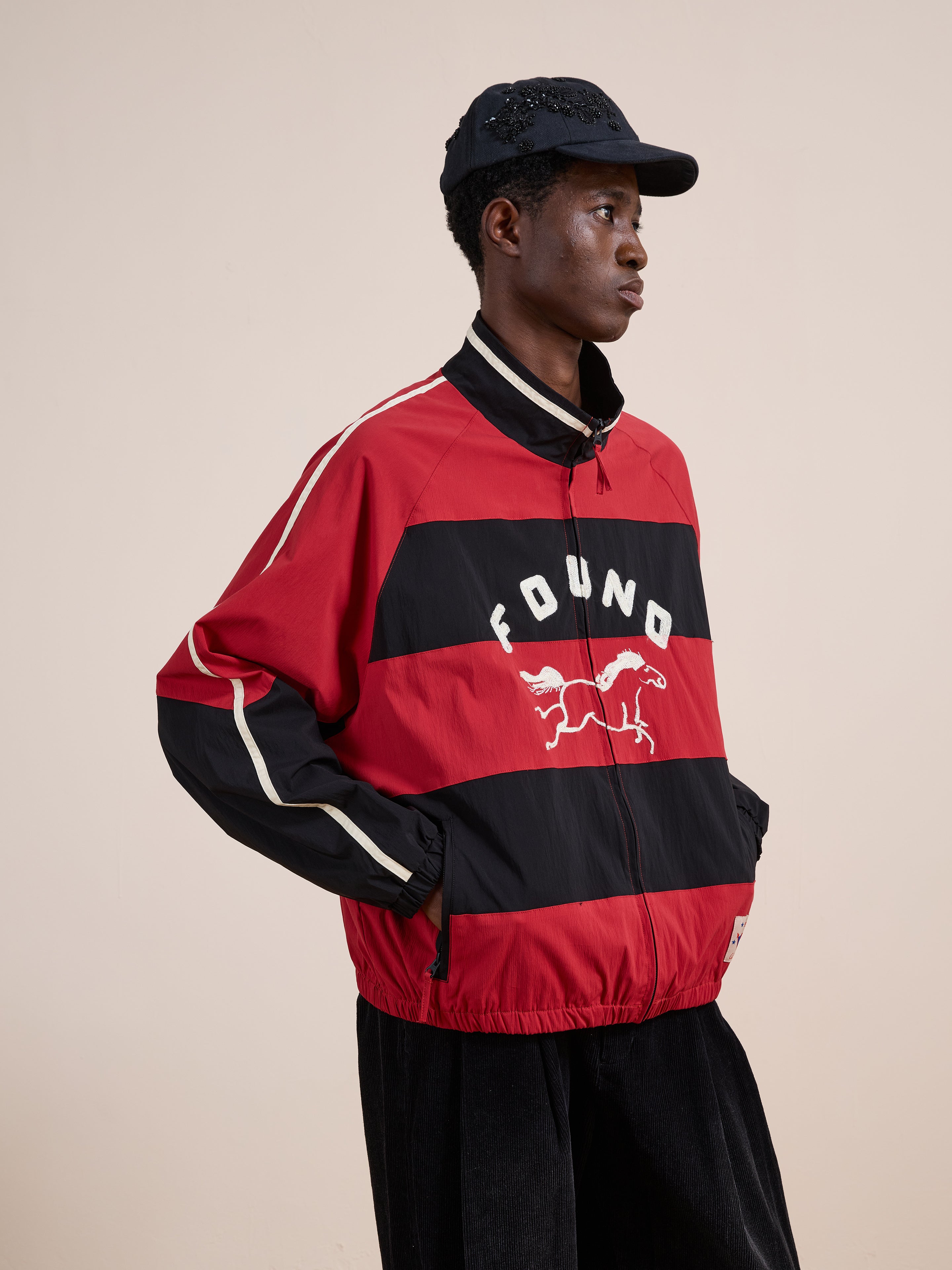 A person wearing the FOUND Racing Striped Windbreaker, featuring vintage sportswear-inspired red and black stripes and a horse graphic, pairs it with a black cap against a plain beige background.