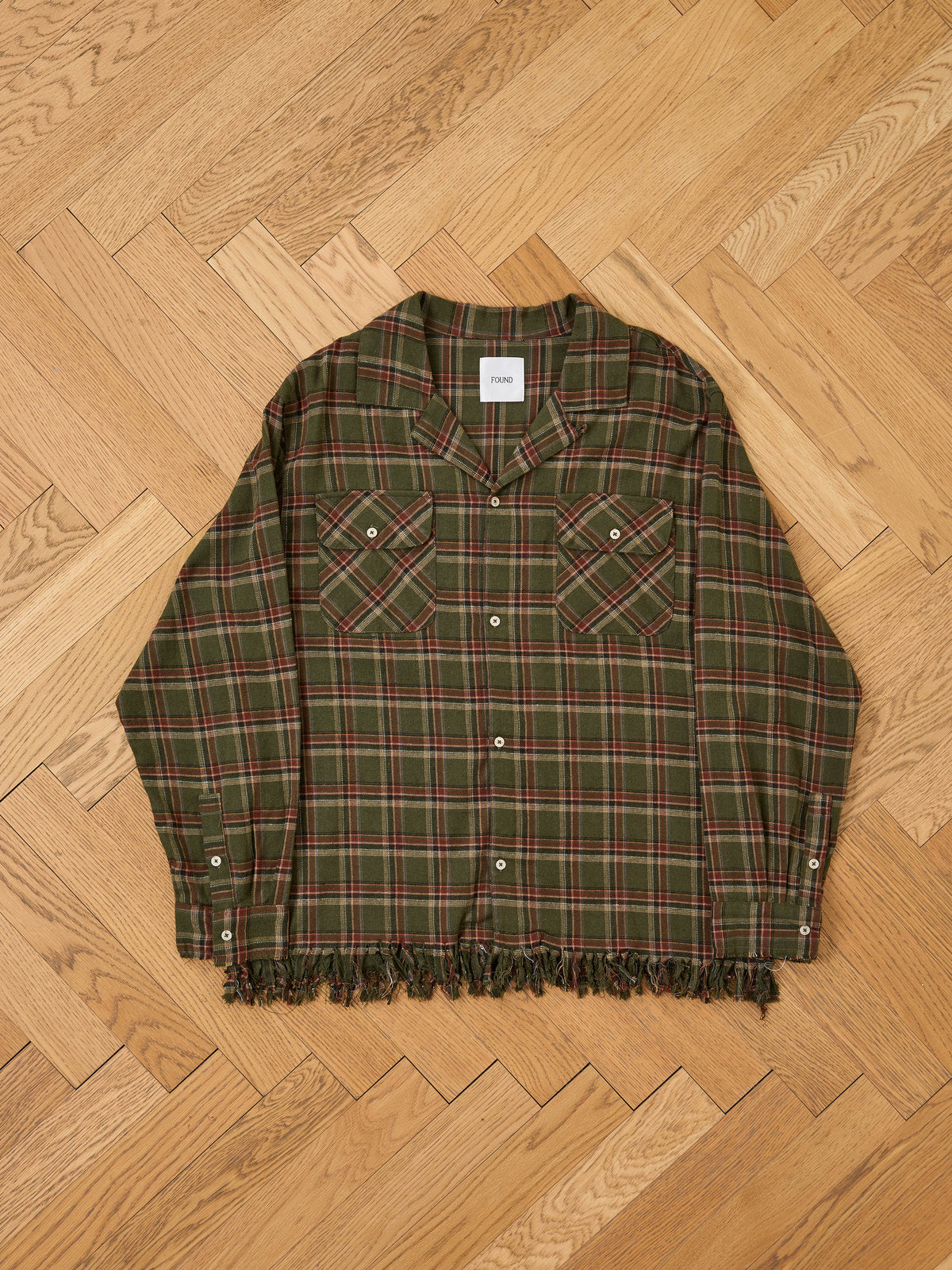 Raw Cut Fringe Plaid Camp Shirt by FOUND in green and red plaid with two chest pockets and a frayed hem, displayed on a light wood herringbone floor.