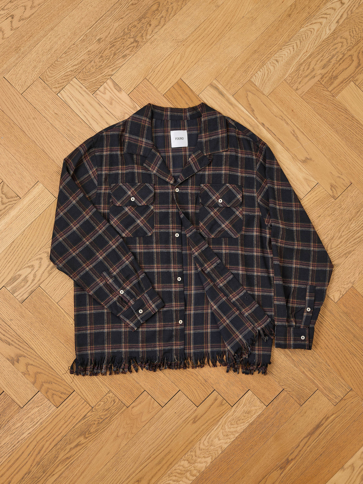 The Raw Cut Fringe Plaid Camp Shirt by FOUND in black and brown plaid with raw cut fringe, button-up front, and two chest pockets is laid flat on a wooden floor.