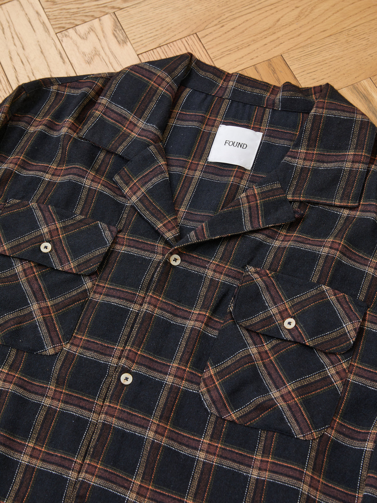 The Raw Cut Fringe Plaid Camp Shirt by FOUND is a unisex overshirt with front pockets, laid flat on a wooden floor. A white “FOUND” tag is visible inside the collar.