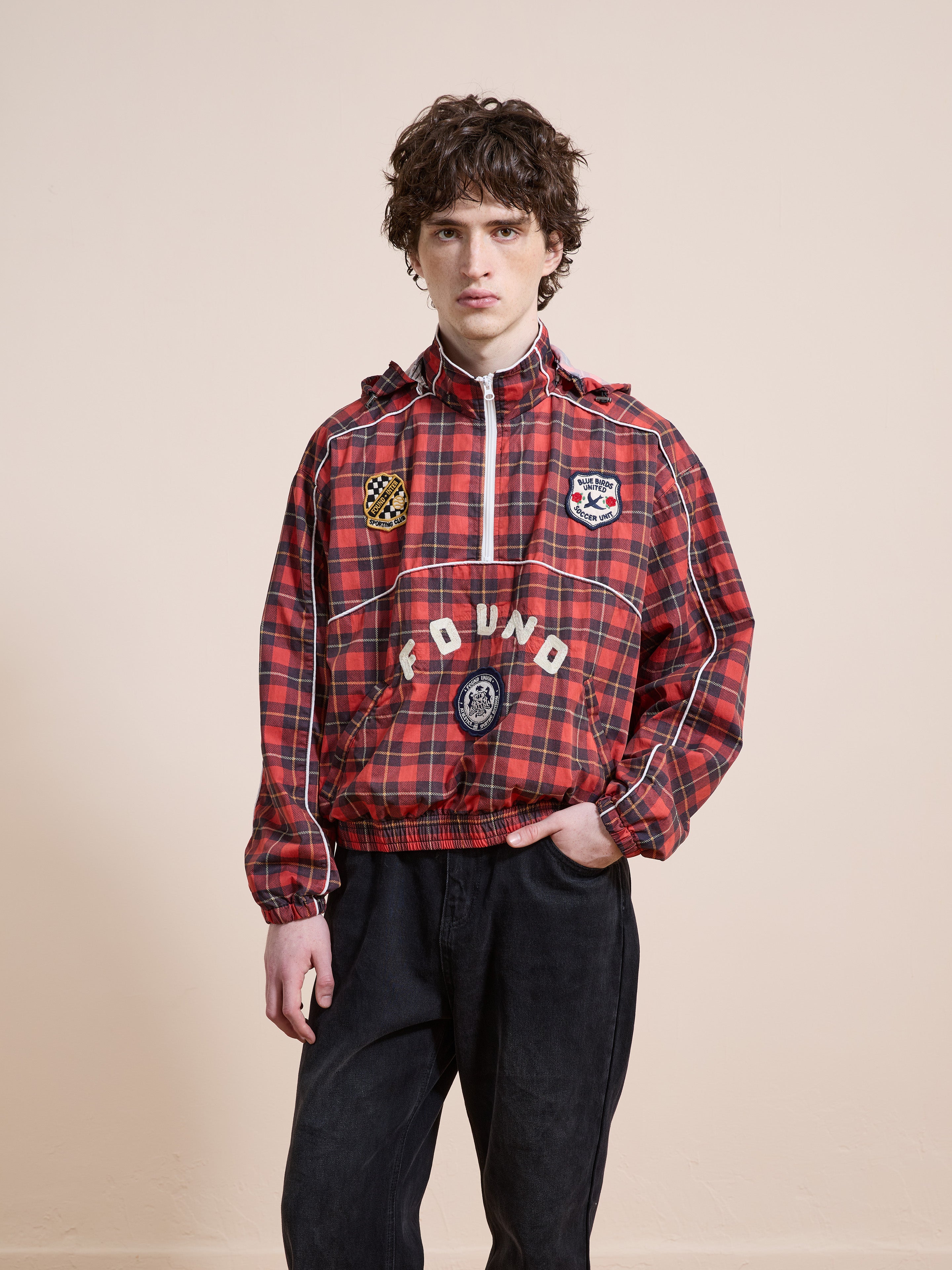 A person stands against a plain background wearing the FOUND Red Plaid Track Jacket, featuring a hood and patches, paired with black pants for a unisex relaxed fit.