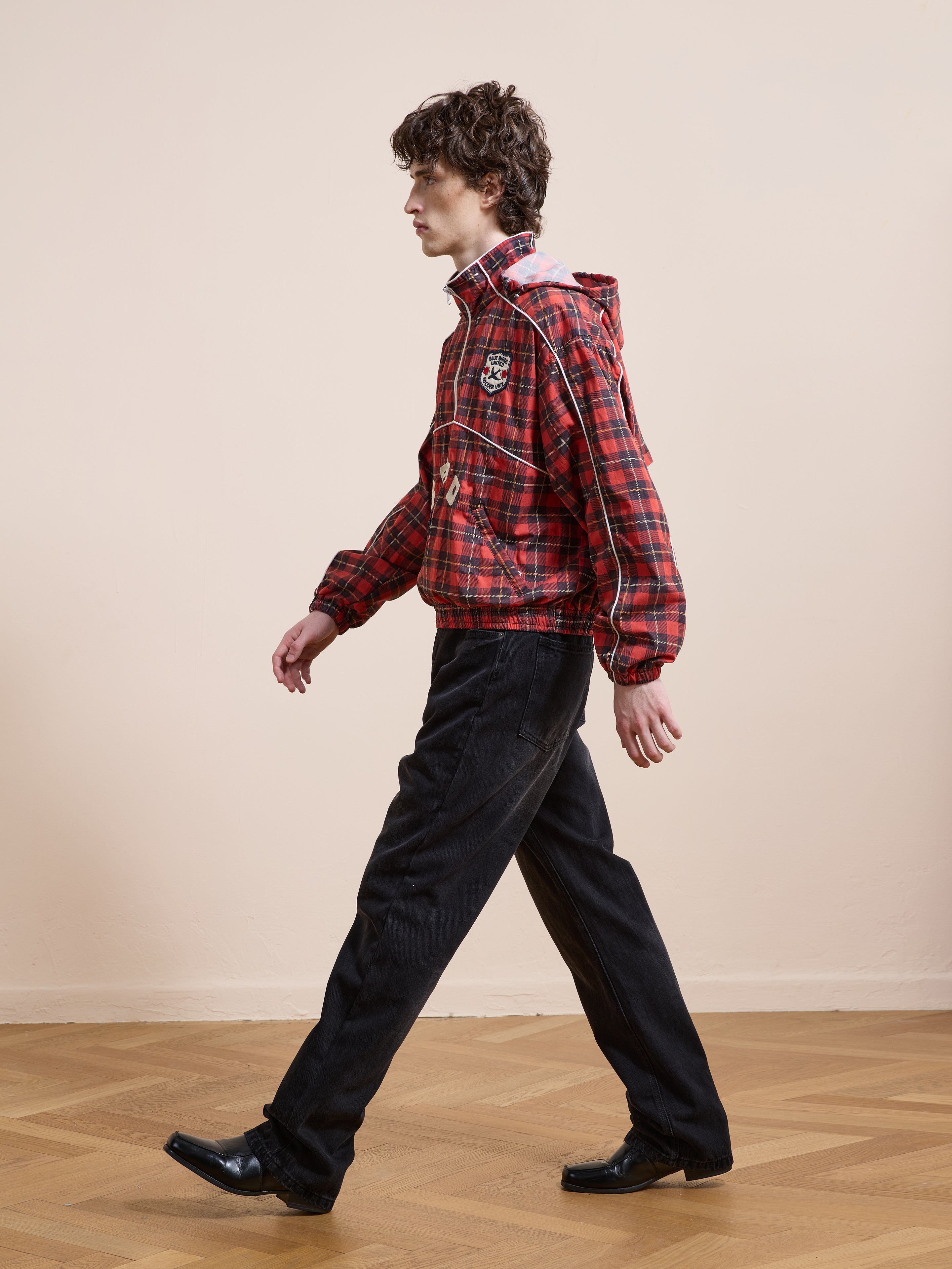 A person wearing the FOUND Red Plaid Track Jacket with a unisex relaxed fit, black jeans, and black shoes walks on a wooden floor in front of a plain beige wall.