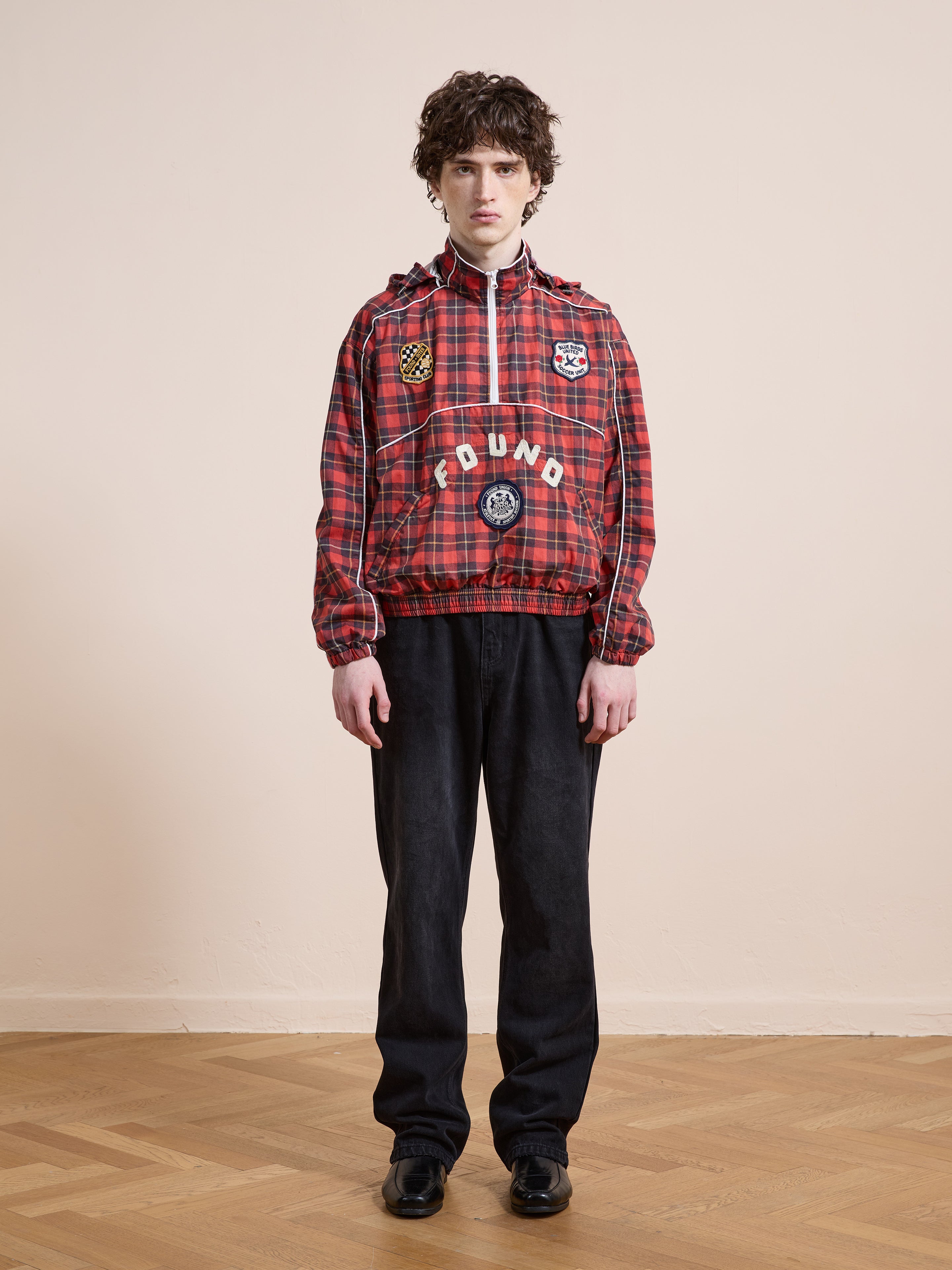 A person stands indoors against a light wall and wooden floor, wearing the FOUND Red Plaid Track Jacket with black pants and black shoes.