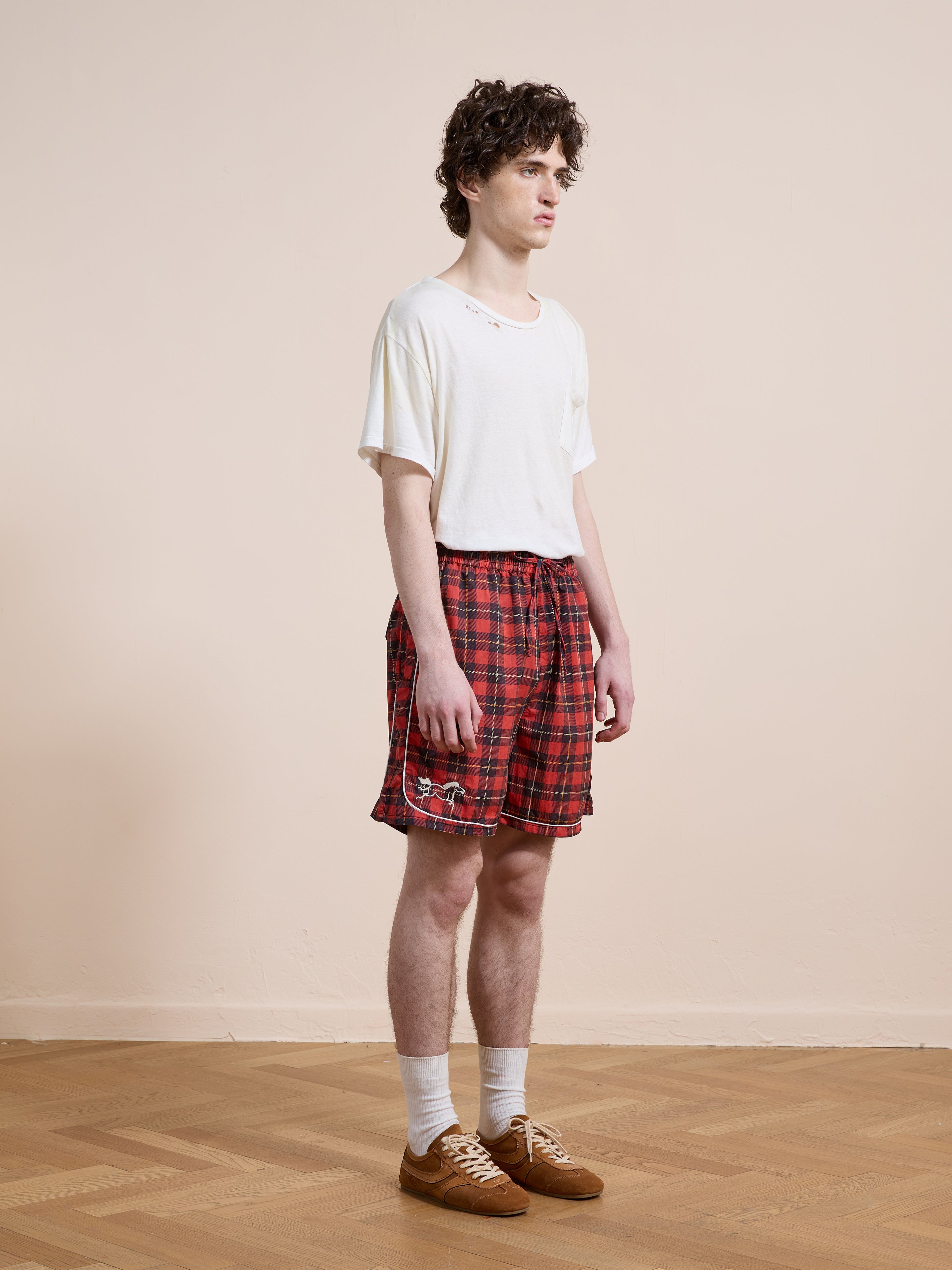 A young man stands indoors in a white t-shirt, Found Red Plaid Track Shorts with a unisex relaxed fit, white socks, and brown sneakers, facing slightly to the side against a plain beige wall.