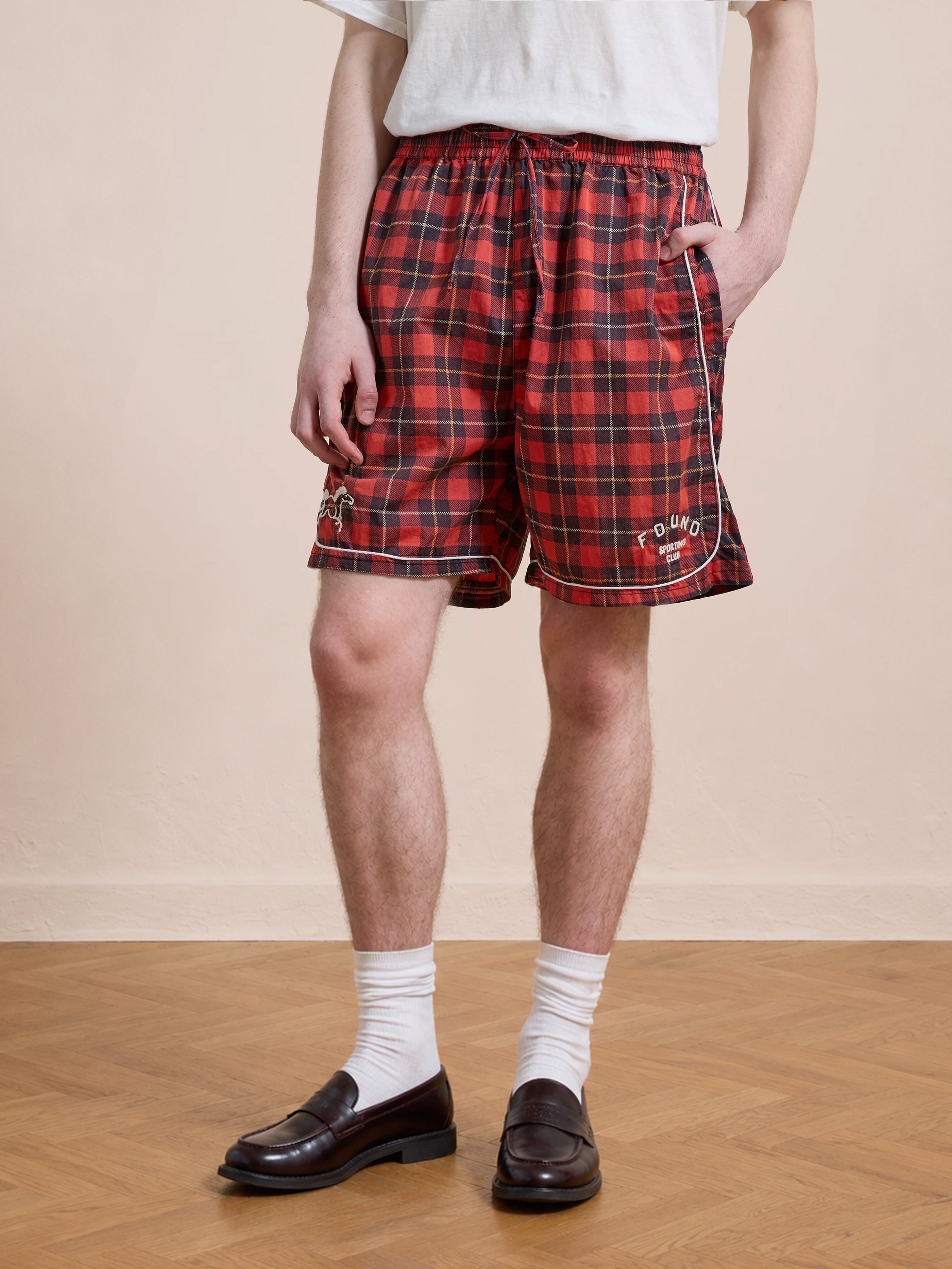 A person wearing a white t-shirt, Found Red Plaid Track Shorts, white socks, and black loafers stands on a wooden floor against a plain beige wall.