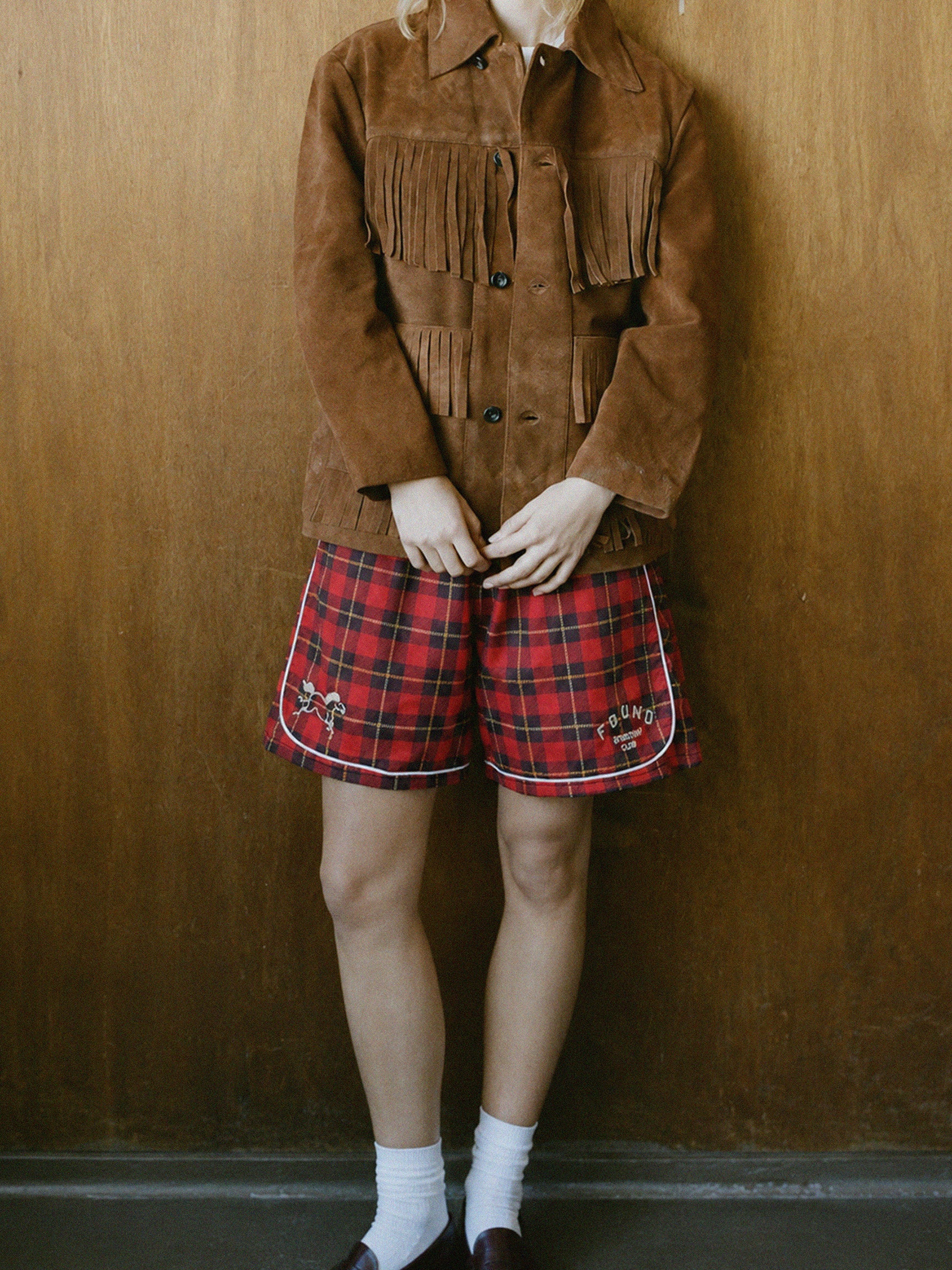 A person stands against a wooden wall wearing Found's Red Plaid Track Shorts, a brown suede fringe jacket, white socks, and dark shoes; their head is not visible.