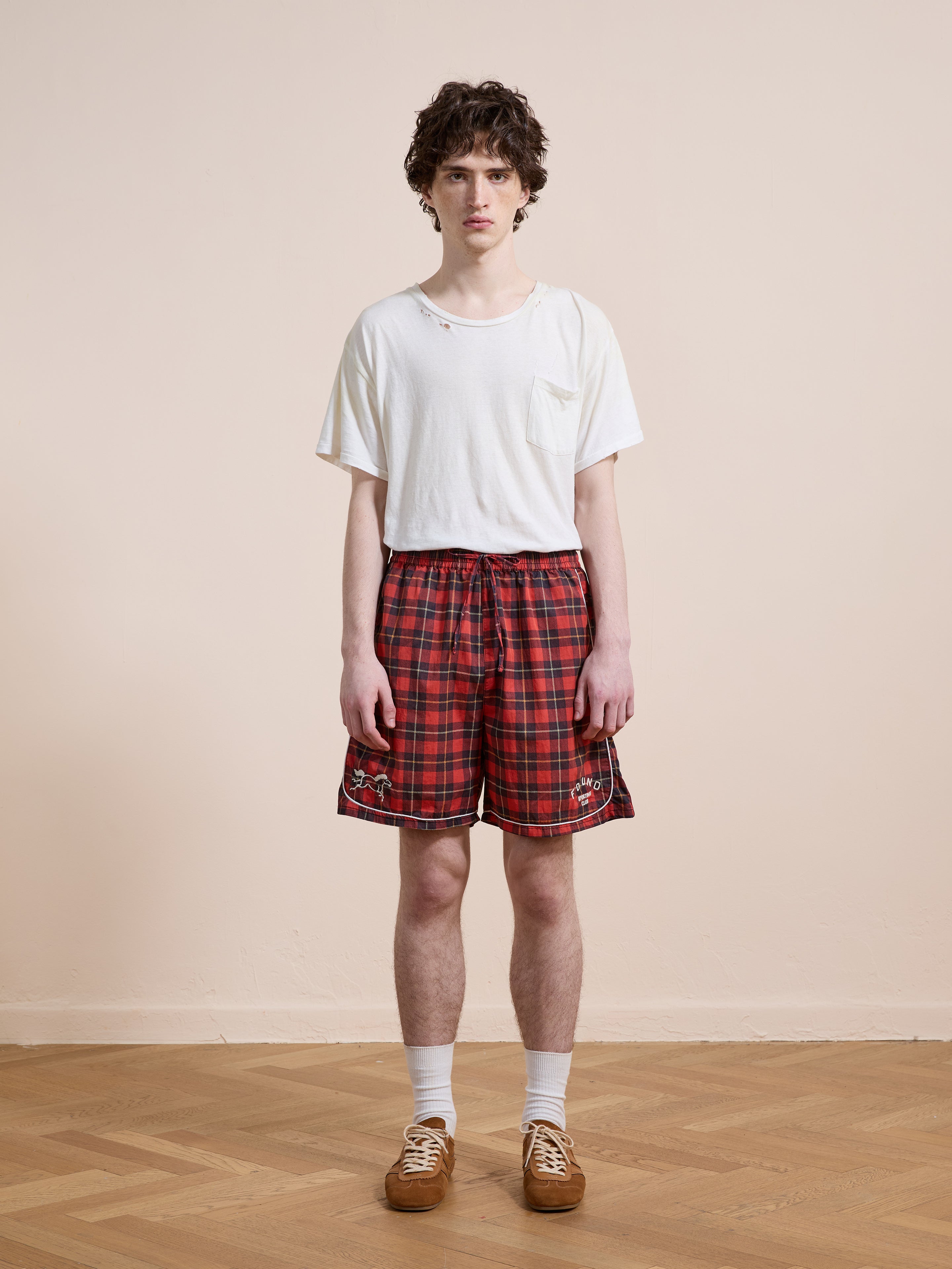 A person stands indoors on a wooden floor, wearing a white t-shirt and Found's Red Plaid Track Shorts with white socks and brown shoes, against a plain beige wall.