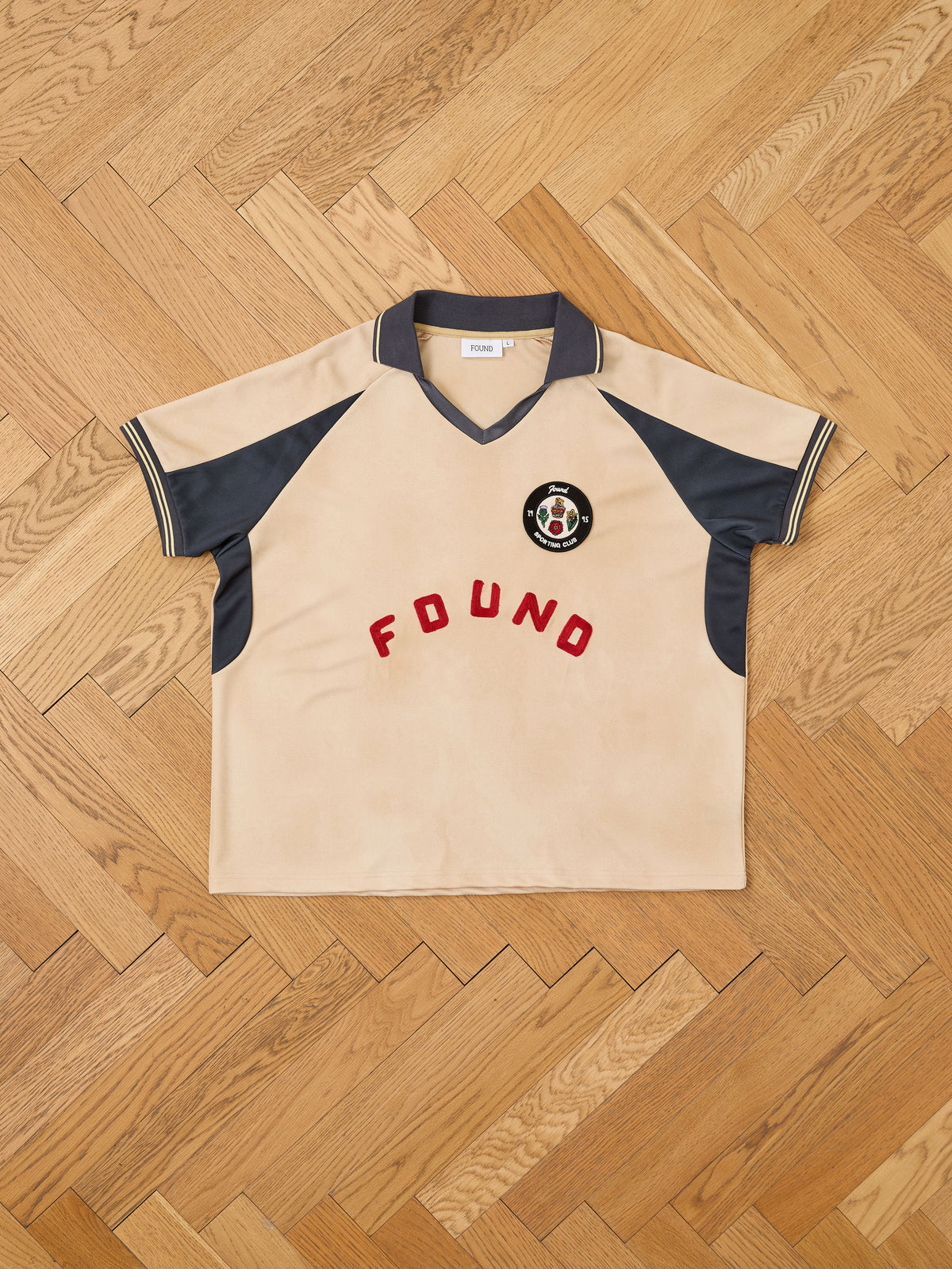 The Rosewood Jersey by FOUND is a beige and navy short-sleeve shirt featuring the “FOUND” logo and a circular chest patch, styled after a vintage football jersey and displayed flat on a wooden parquet floor.