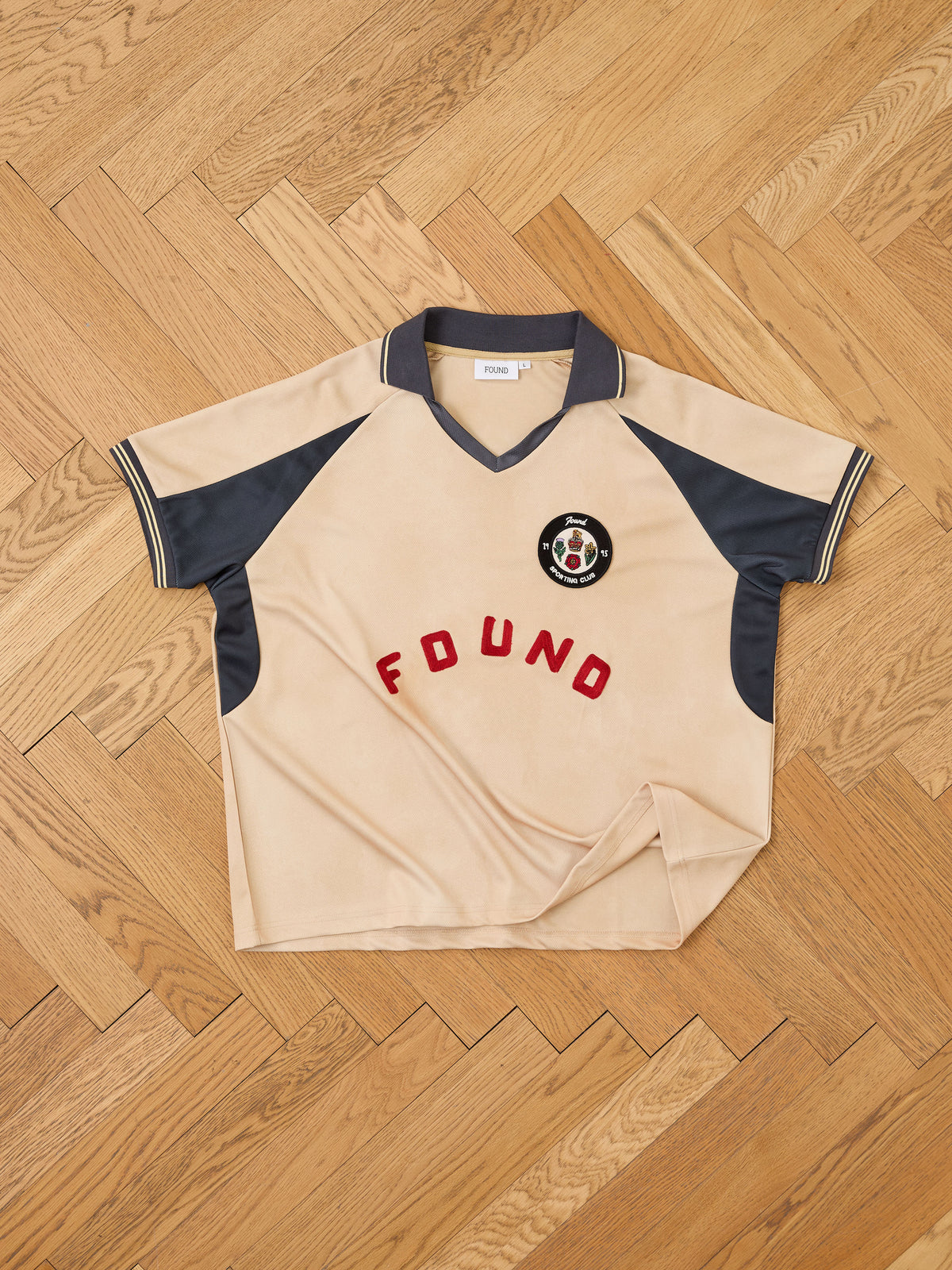 The Rosewood Jersey by FOUND is a beige and navy short-sleeve polo shirt, inspired by vintage football jersey styling, displayed on a parquet floor. It features the word “FOUND” and a circular chest patch with a bear graphic.