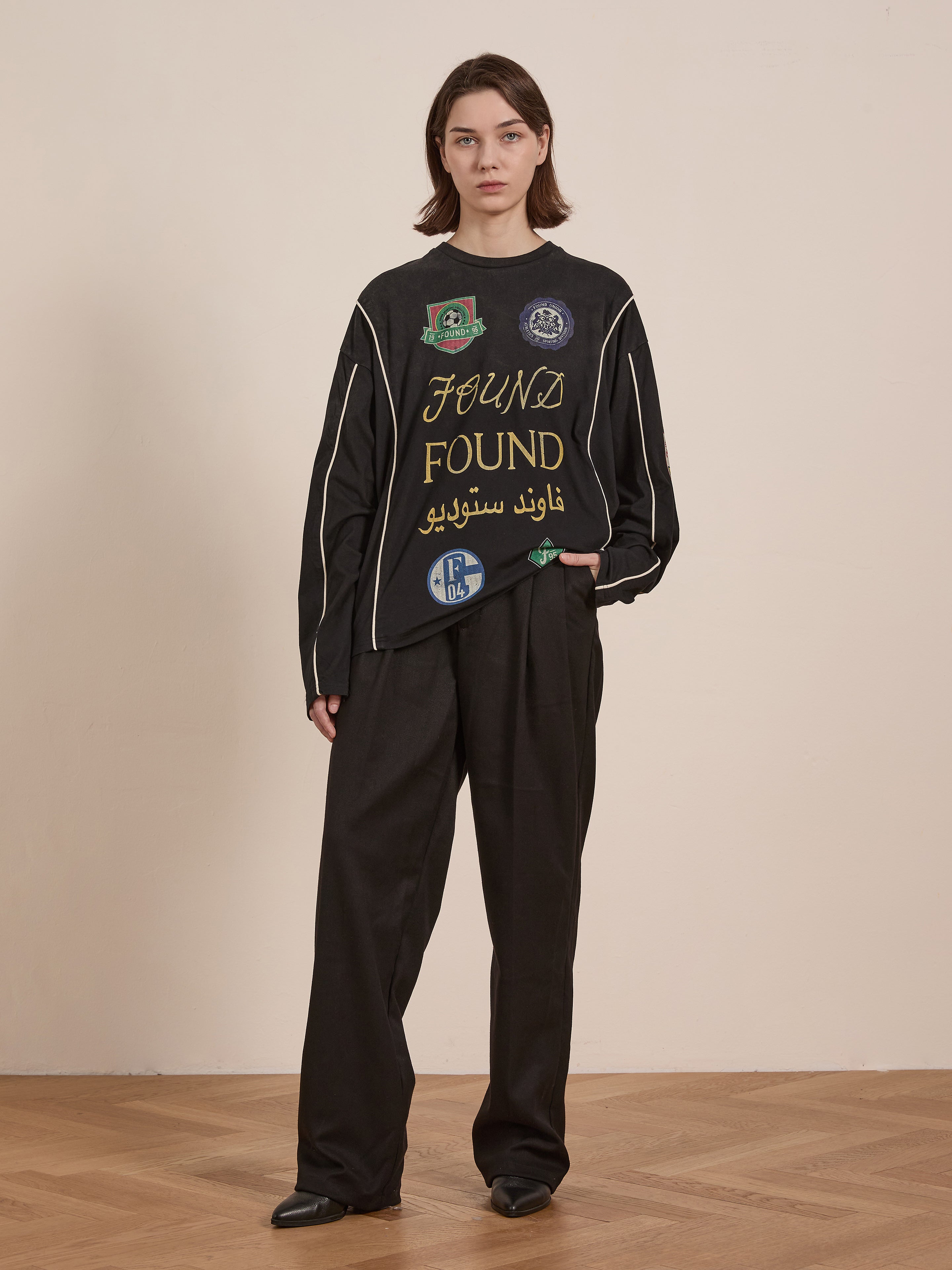 A person wearing FOUND's Multi Language LS Jersey, a unisex black outfit with embroidered text and symbols and a retro silhouette, stands on a wooden floor against a plain wall, echoing the simplicity of a classic football jersey.