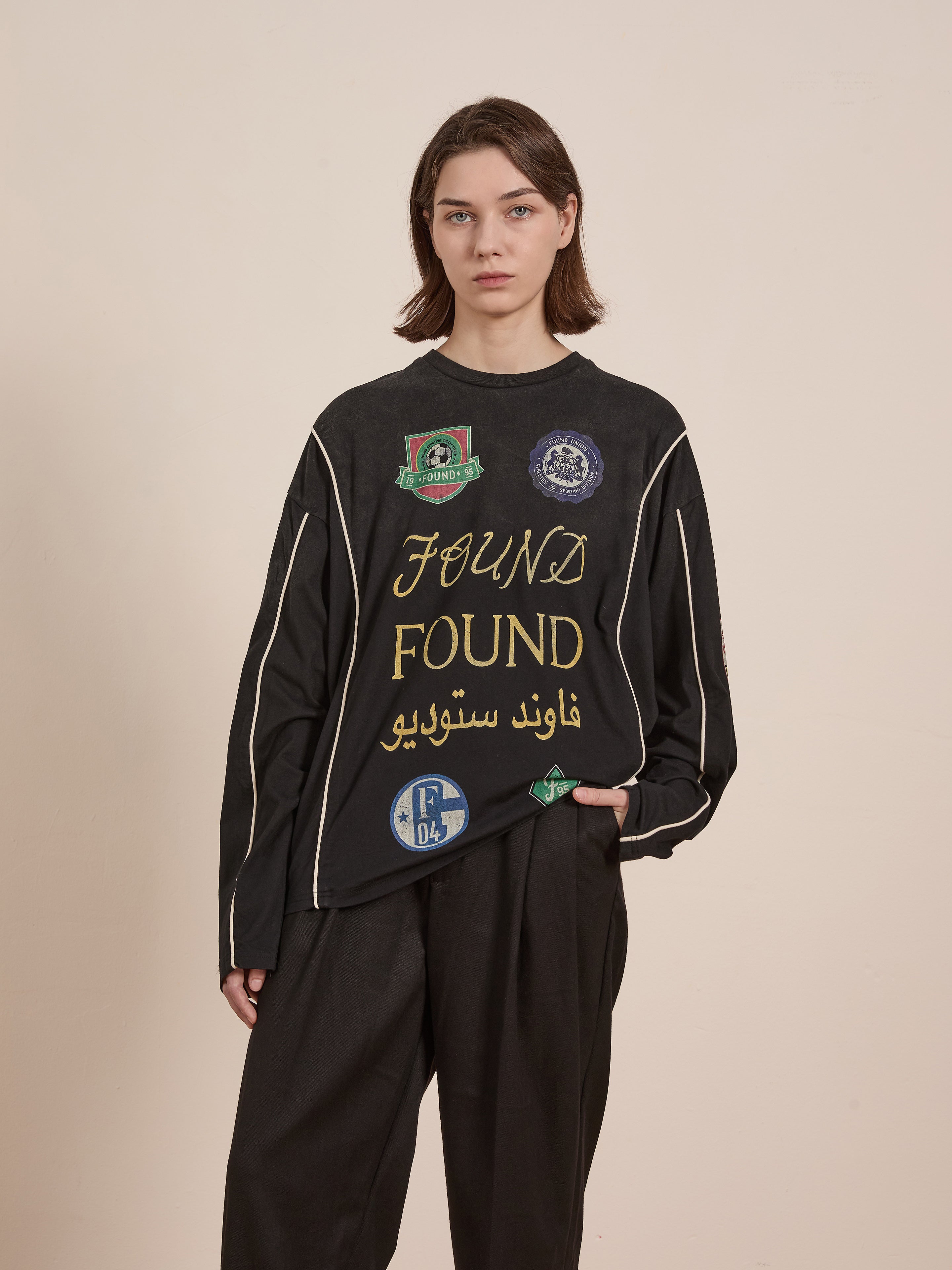 A woman models FOUND's Multi Language LS Jersey— a retro black long-sleeve shirt featuring "FOUND" and logos in English and Arabic, paired with loose black pants, all set against a plain beige backdrop.