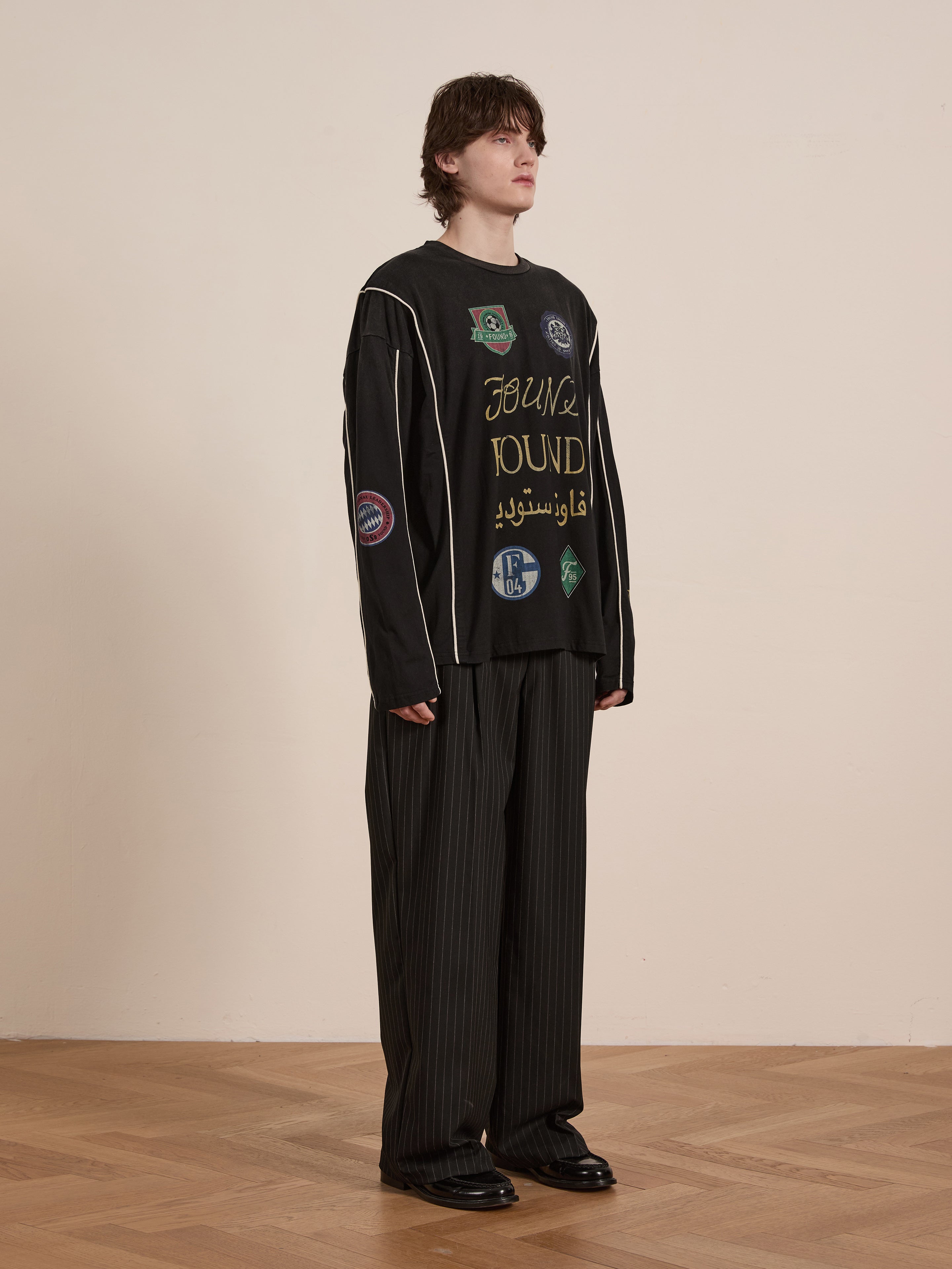 A person with shoulder-length hair sports the FOUND brand's Multi Language LS Jersey, covered in various patches and text, paired with pinstripe pants and black shoes. Standing on a wooden floor against a beige wall, this unisex outfit effortlessly captures a retro silhouette.