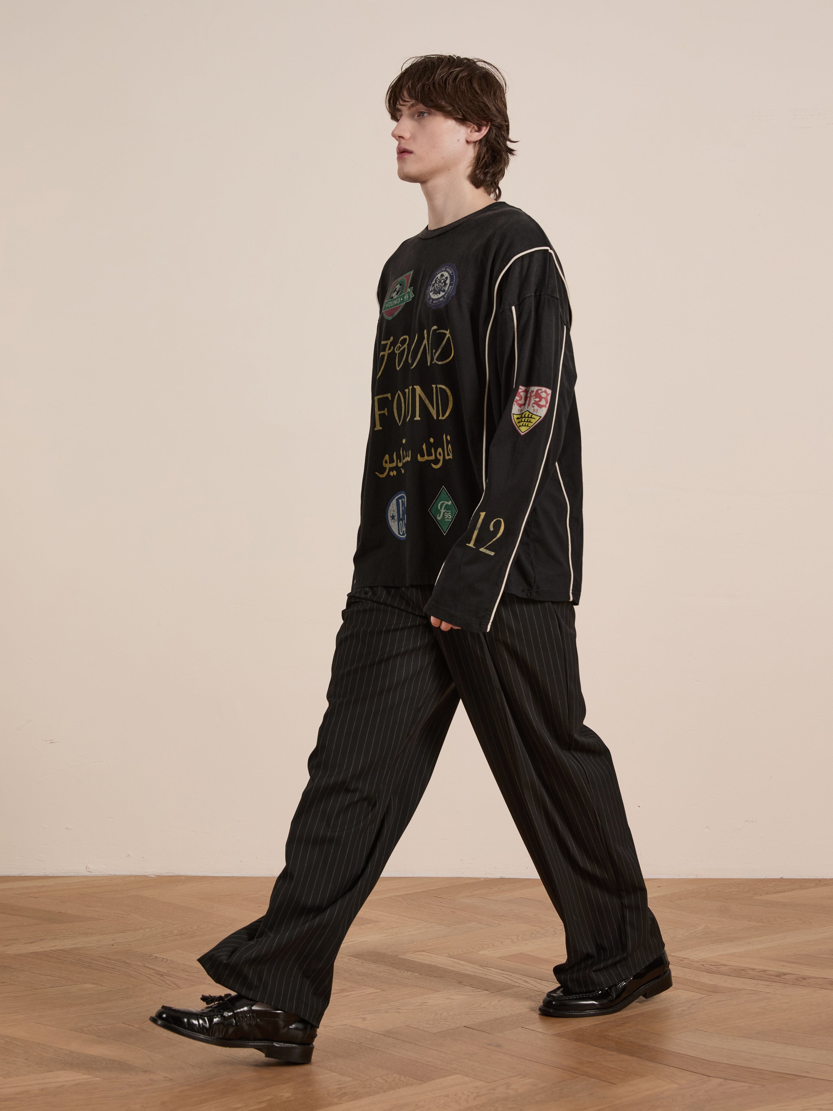 A person walks on a wooden floor, dressed in FOUND's Multi Language LS Jersey, which features colorful patches with a retro silhouette. The look is completed with black pinstripe pants and shiny black shoes against a beige wall.