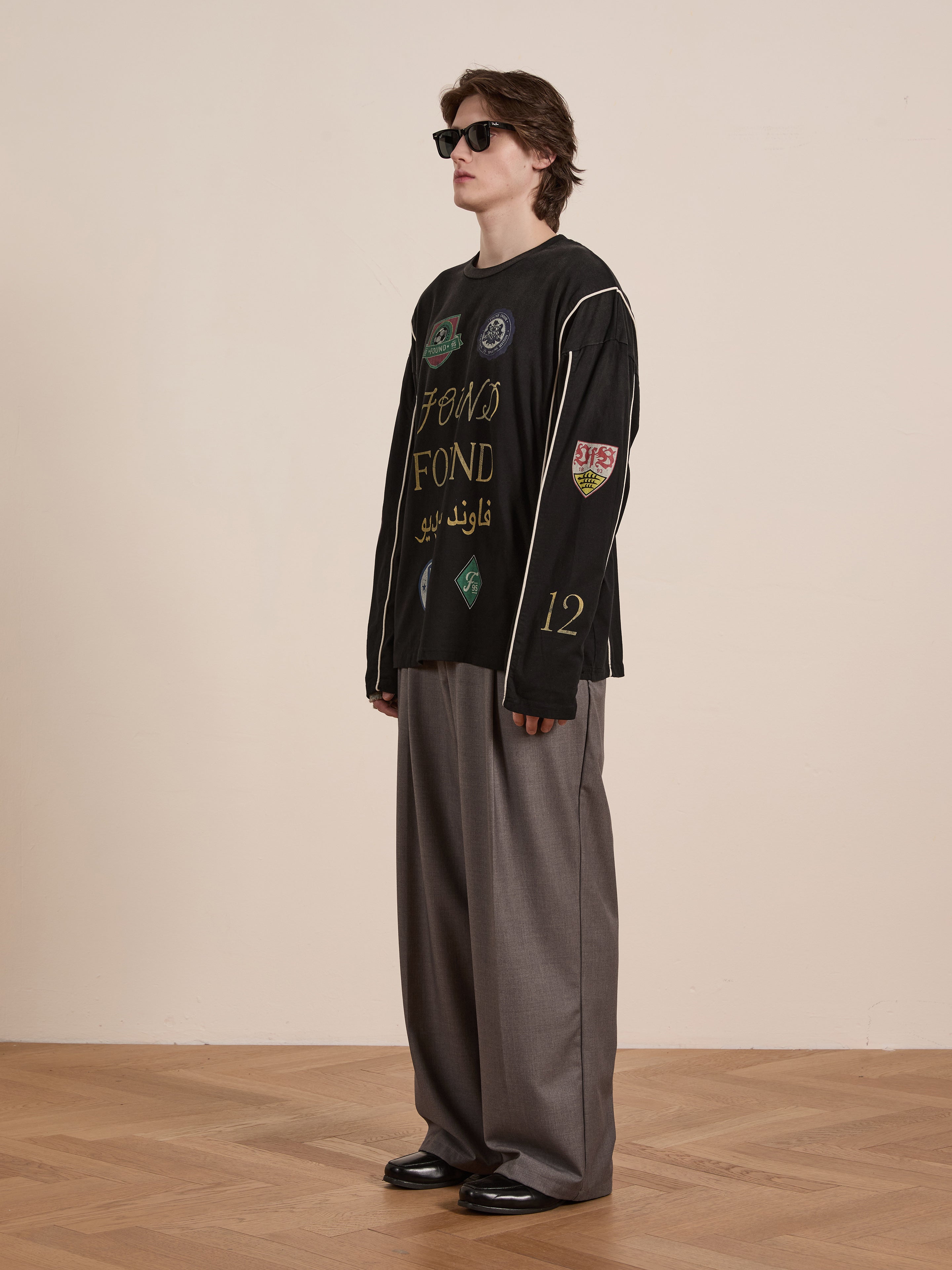 Wearing FOUND's Multi Language LS Jersey, a person stands confidently on a wooden floor. Their gray wide-leg pants create a retro silhouette, capturing the essence of unisex casual wear against a plain background. They're accessorized with sunglasses to complete the look.