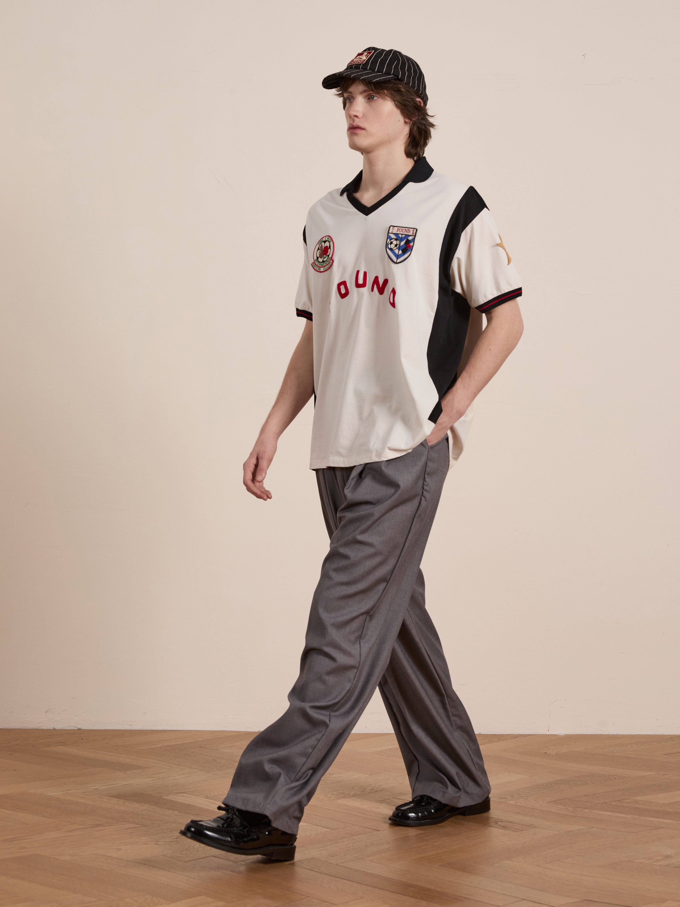 Wearing a Union Soccer Jersey by FOUND, paired with gray pants and black shoes, a person with a striped cap walks indoors on a wooden floor, blending vintage football charm with contemporary style.