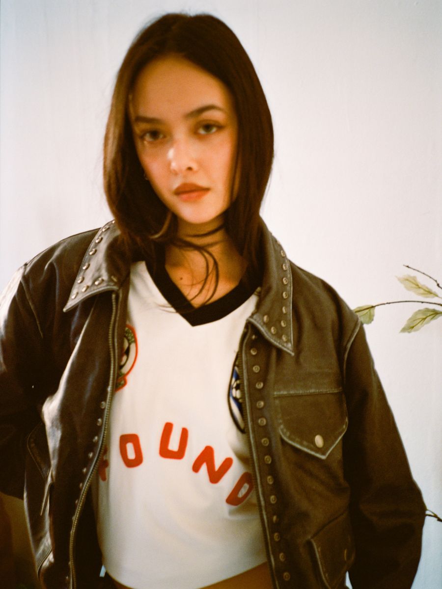 A person with long dark hair wears the FOUND Studded Leather Pocket Jacket over a white shirt with red text, standing against a light-colored background.