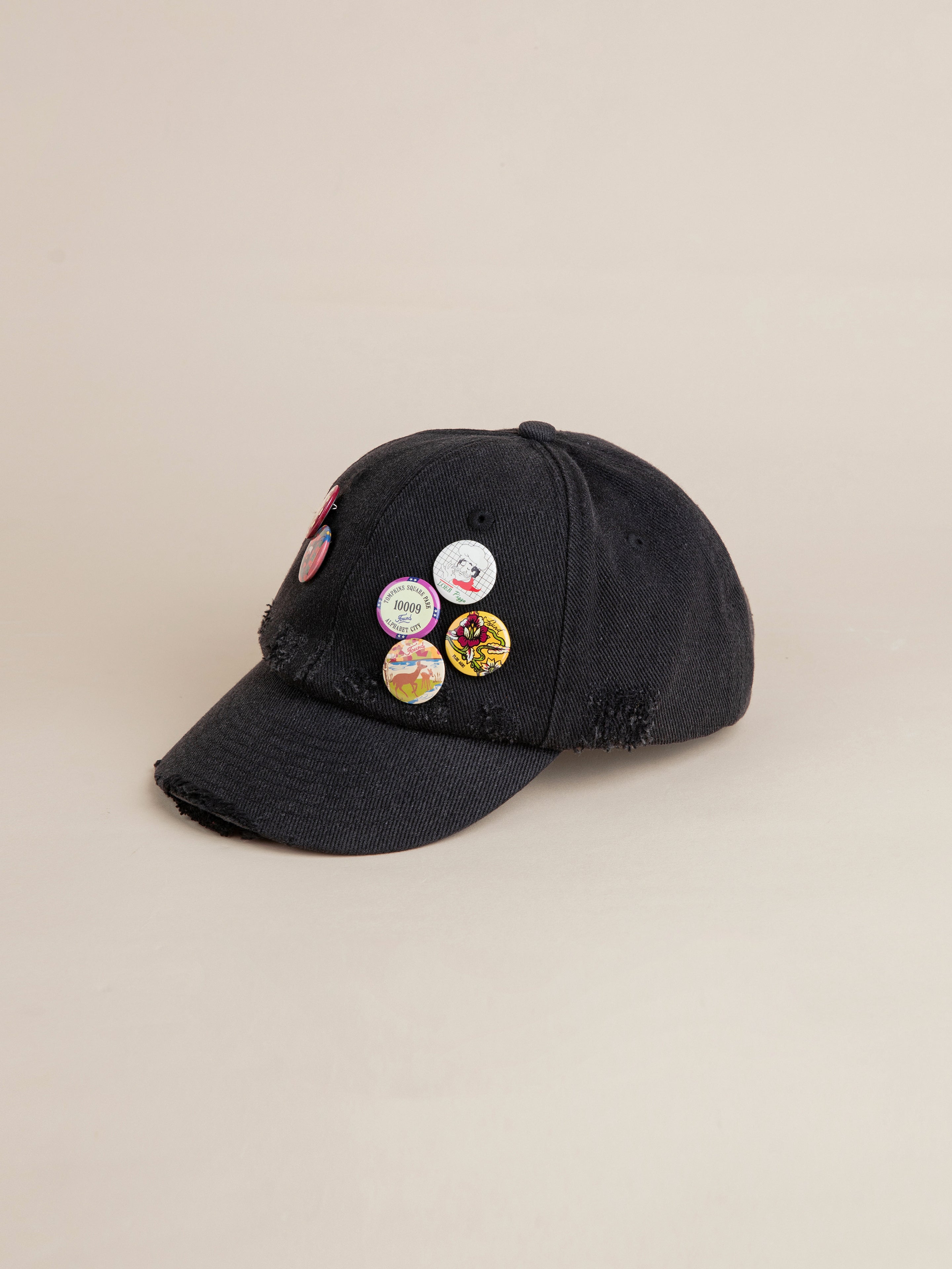 The Distressed Pin Cap by FOUND, a black cotton cap adorned with multiple vintage-inspired pins on the front and side, is displayed against a plain background.