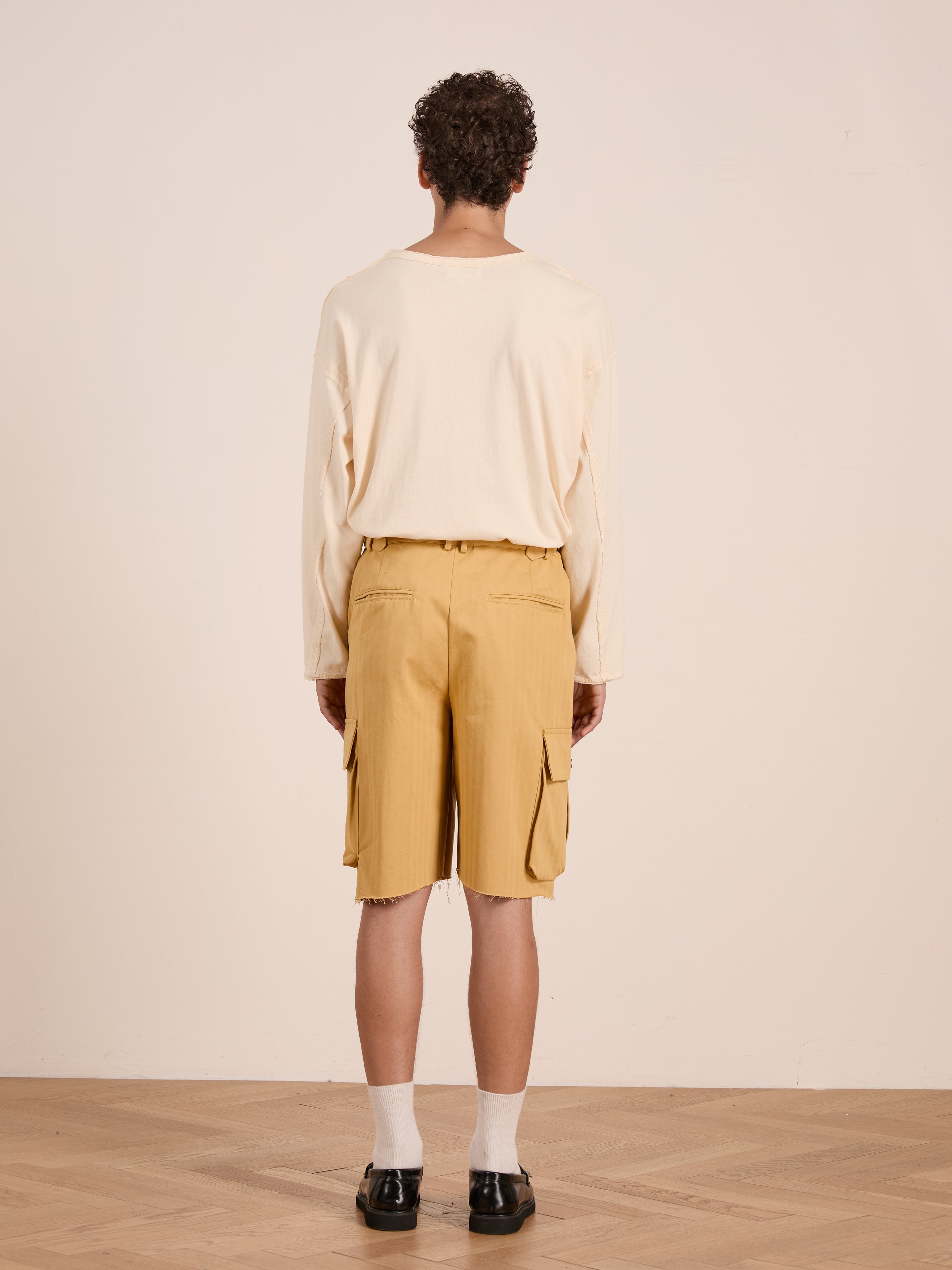 A person stands facing away on a wooden floor against a plain light wall, wearing a long-sleeve beige shirt, white socks, black shoes, and FOUND Sunlight Twill Cargo Shorts in yellow.