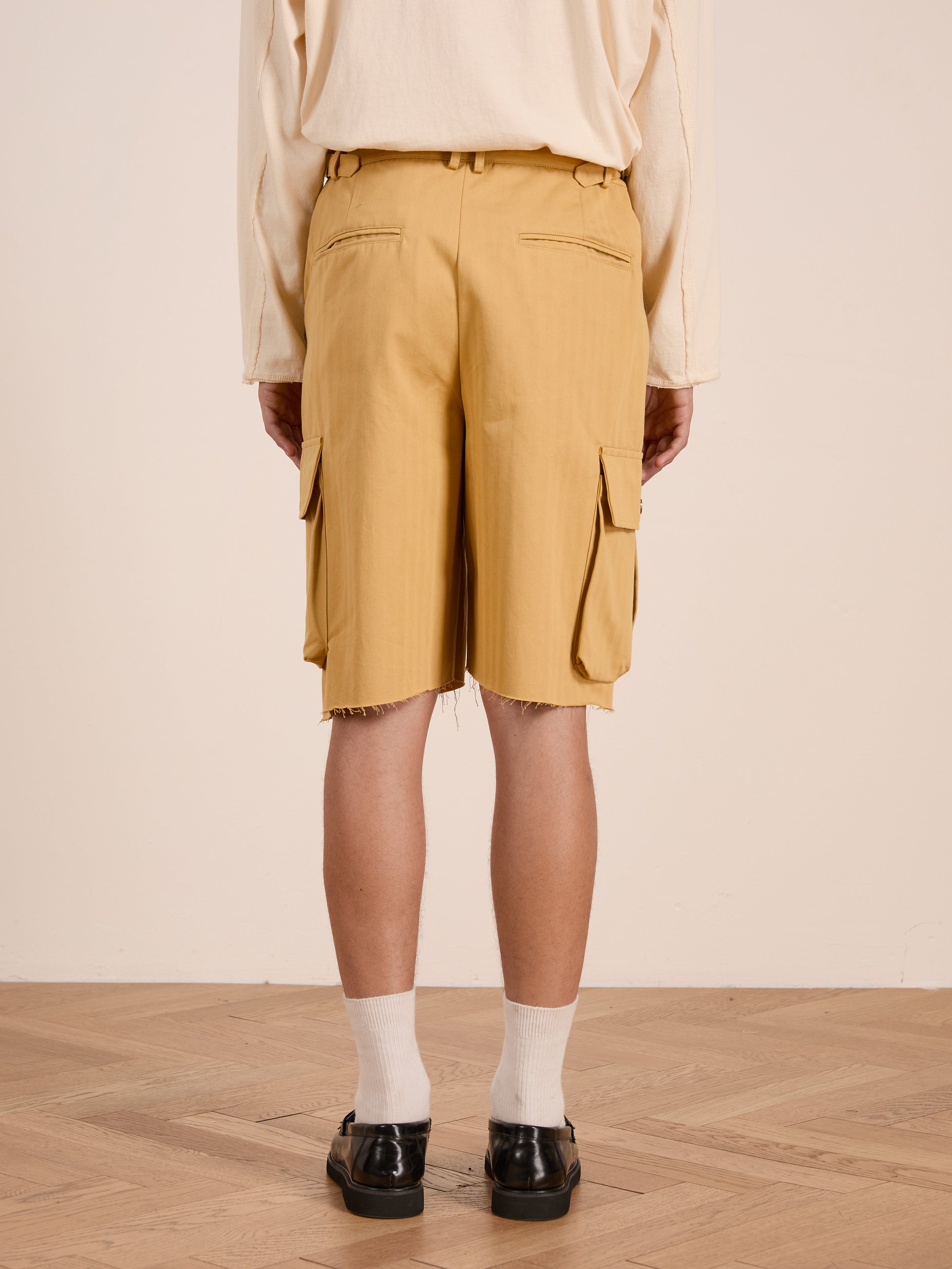 A person stands indoors on a wooden floor, shown from the back, wearing a cream long-sleeve shirt, FOUND Sunlight Twill Cargo Shorts with raw hems in yellow herringbone, white socks, and black shoes.