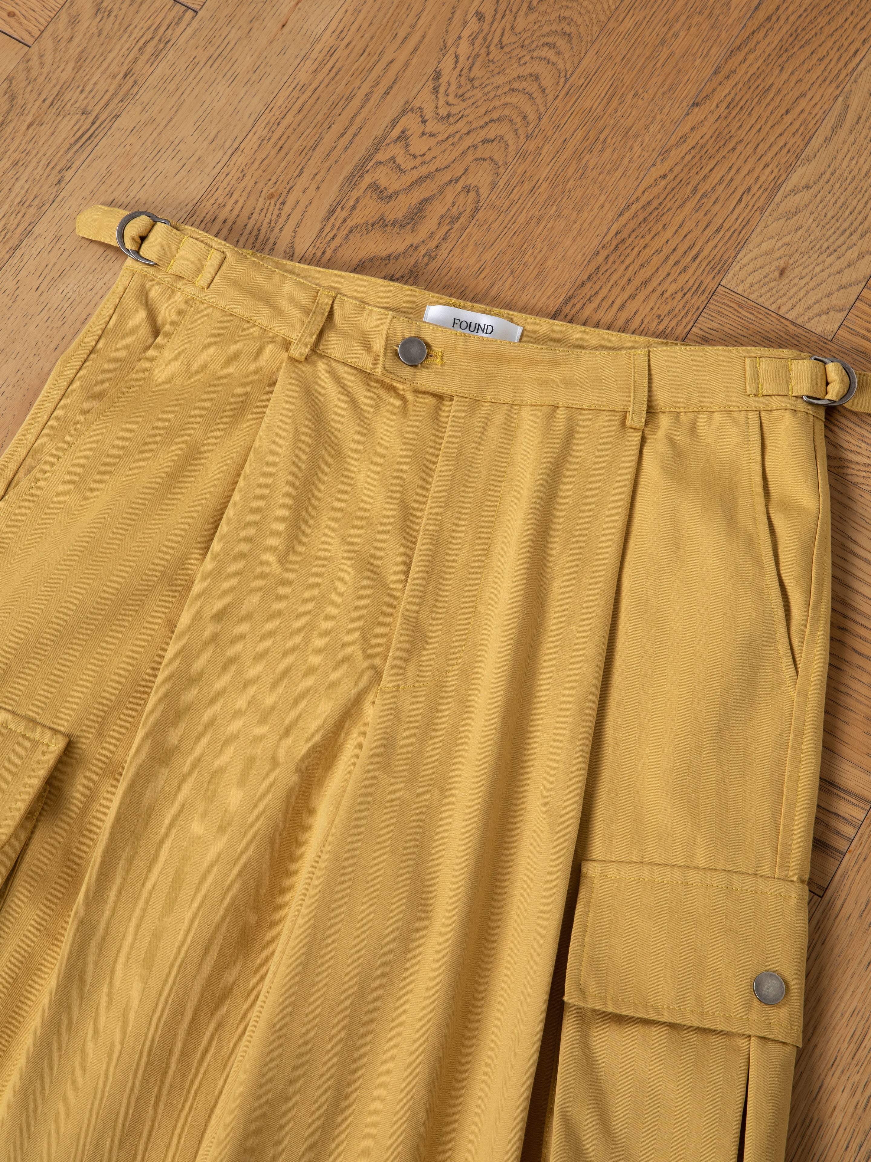 A close-up of FOUND Sunlight Twill Cargo Shorts—unisex, mustard yellow with pleats and flap pockets—laid flat on a wooden floor.
