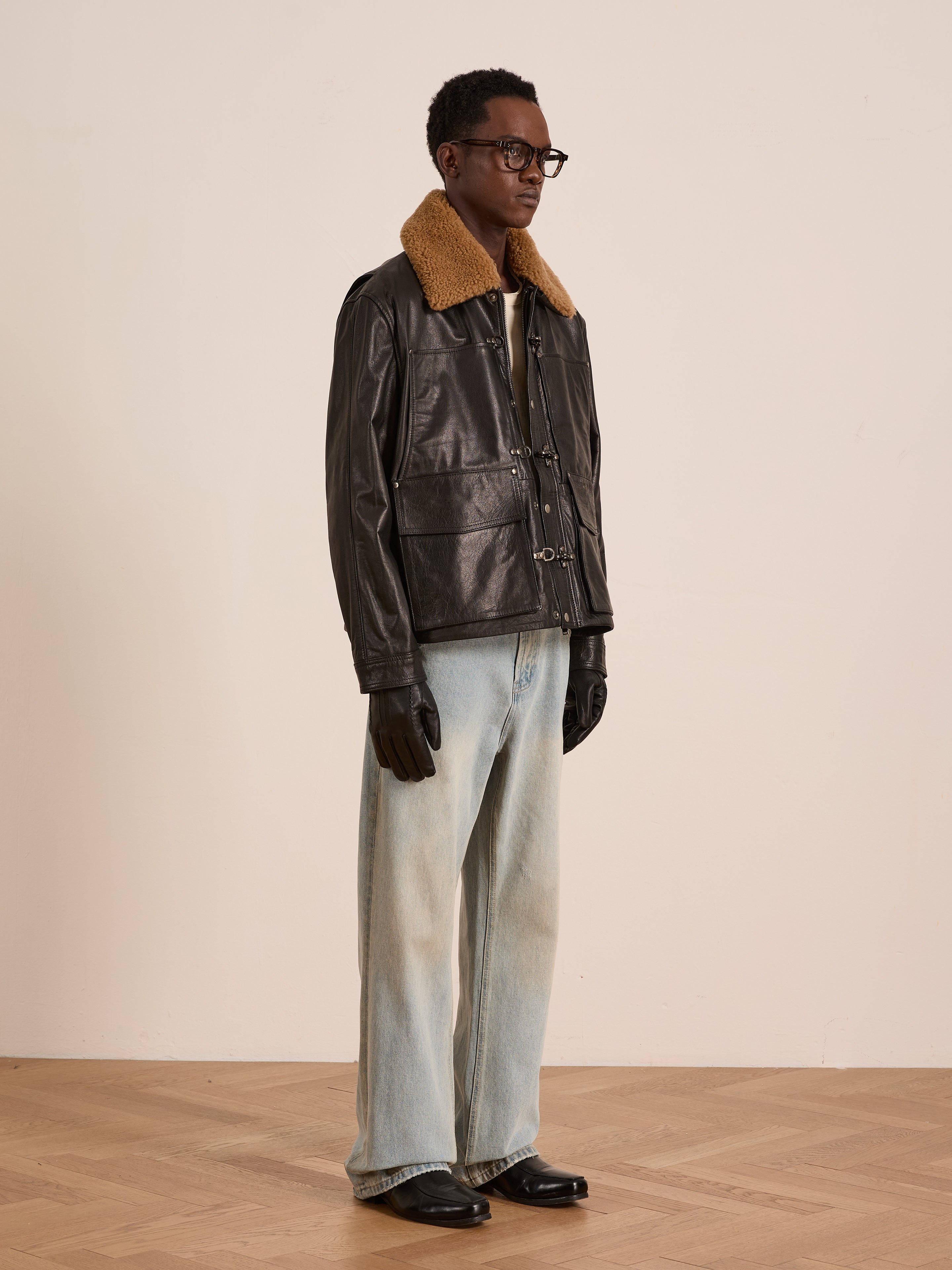 A person in a FOUND Sherpa Collar Leather Utility Jacket, light blue wide-leg jeans, black gloves, and black shoes stands on a wooden floor against a plain background.