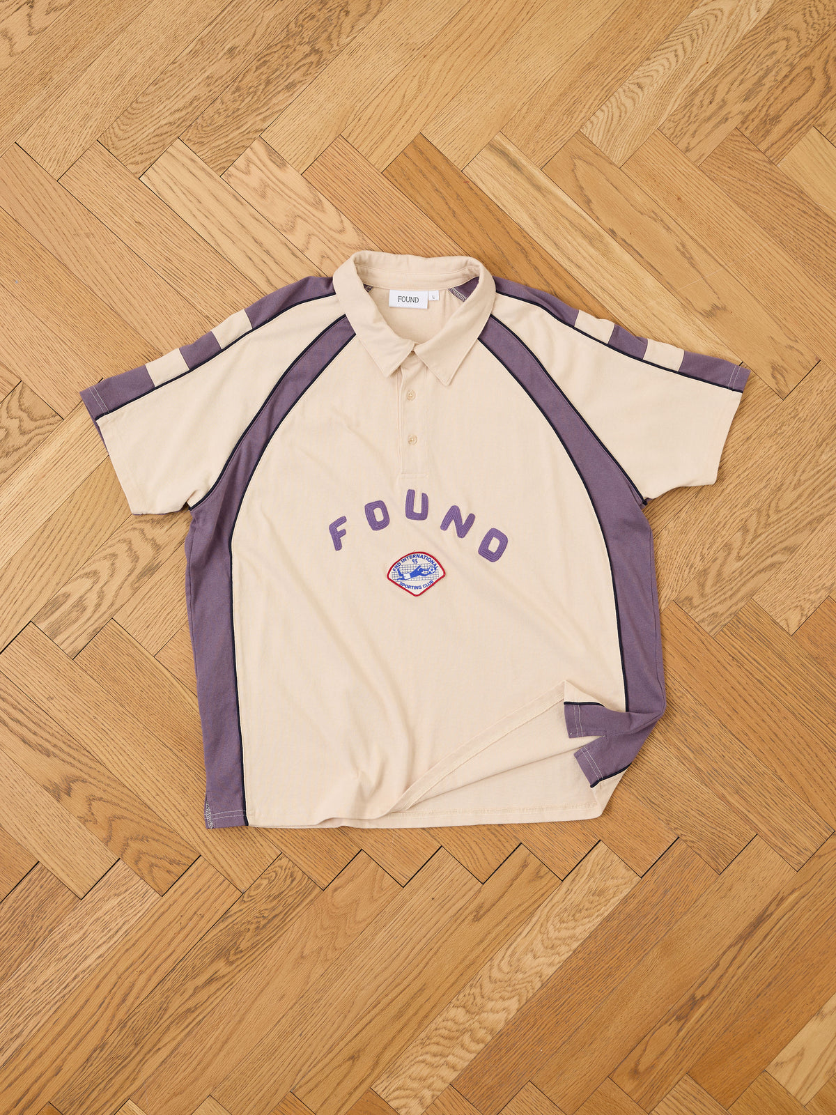 FOUND Short Sleeve Rugby Polo in beige and purple, unisex relaxed fit with front logo patch, shown laid flat on a wooden herringbone floor.