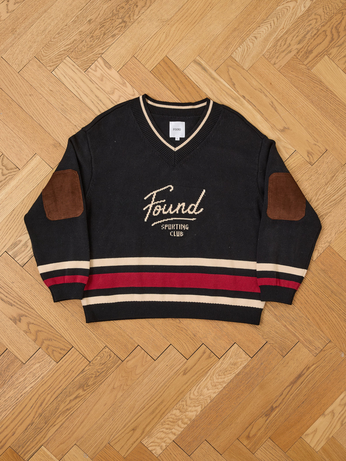FOUND Sport Club Varsity Sweater—unisex, relaxed fit, with beige/red stripes and brown elbow patches, “Found Sporting Club” text, shown laid flat on a wooden floor.