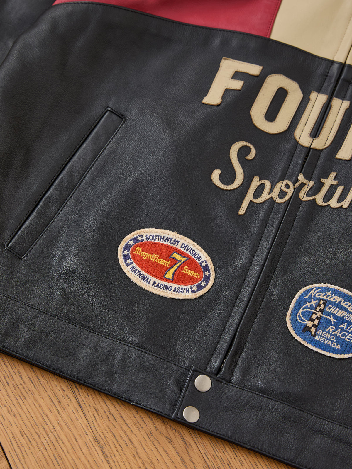 Close-up of the Sport Racer Jacket by FOUND, showcasing embroidered text, racing patches, and a snap button on a wooden surface.
