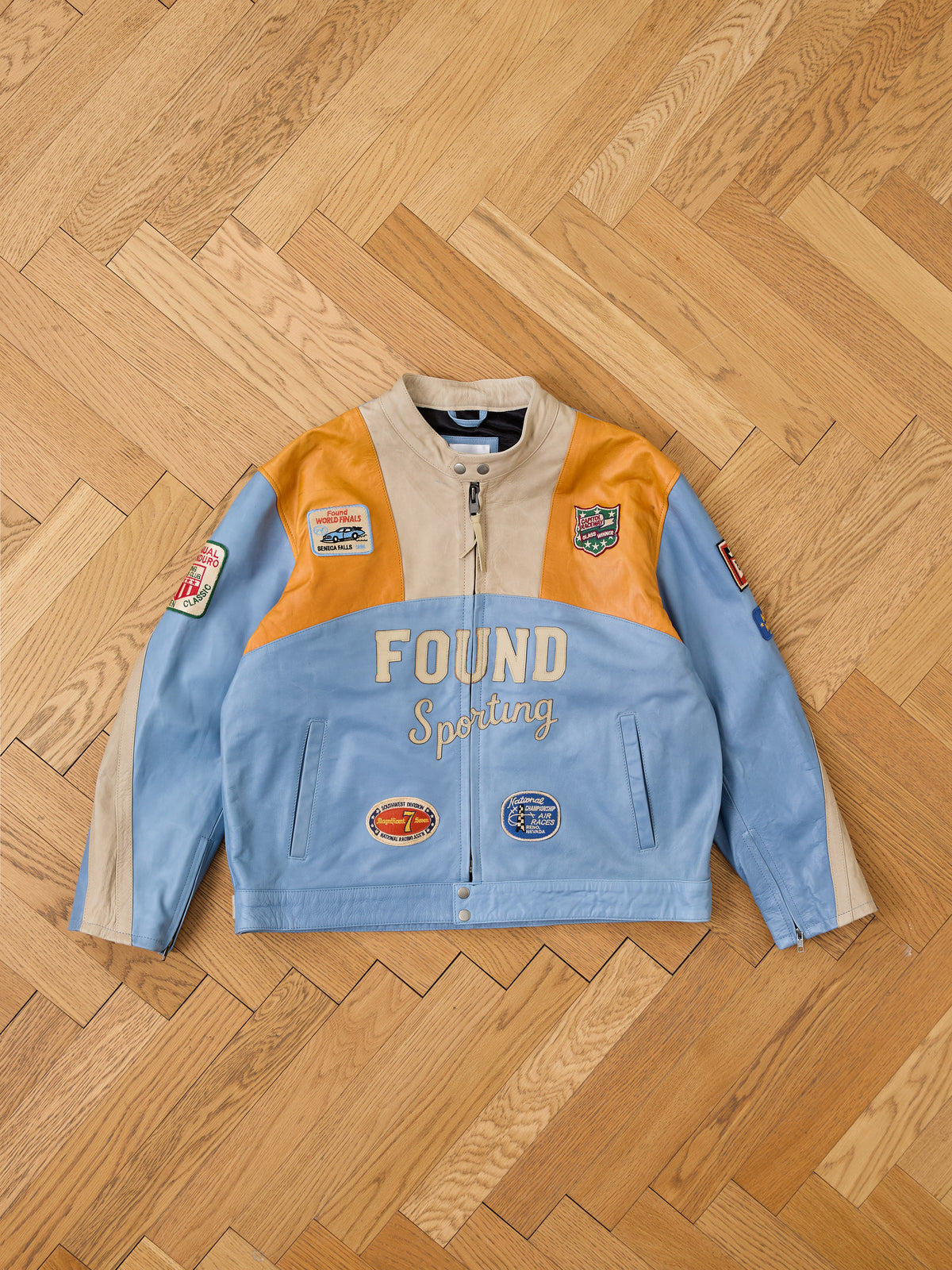 The FOUND Sport Racer Jacket features color-blocked leather, racing patches, and “FOUND Sporting” text, shown on a wood parquet floor.