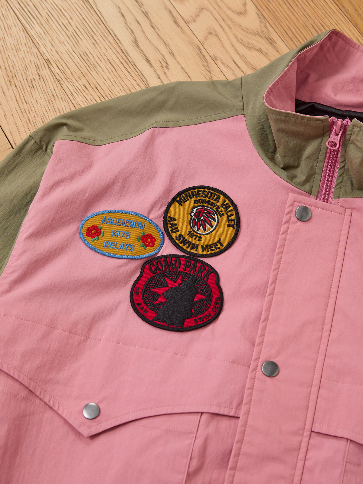 The FOUND Sport Windbreaker Jacket in pink and olive green features three embroidered chest patches and is displayed on a wooden floor.