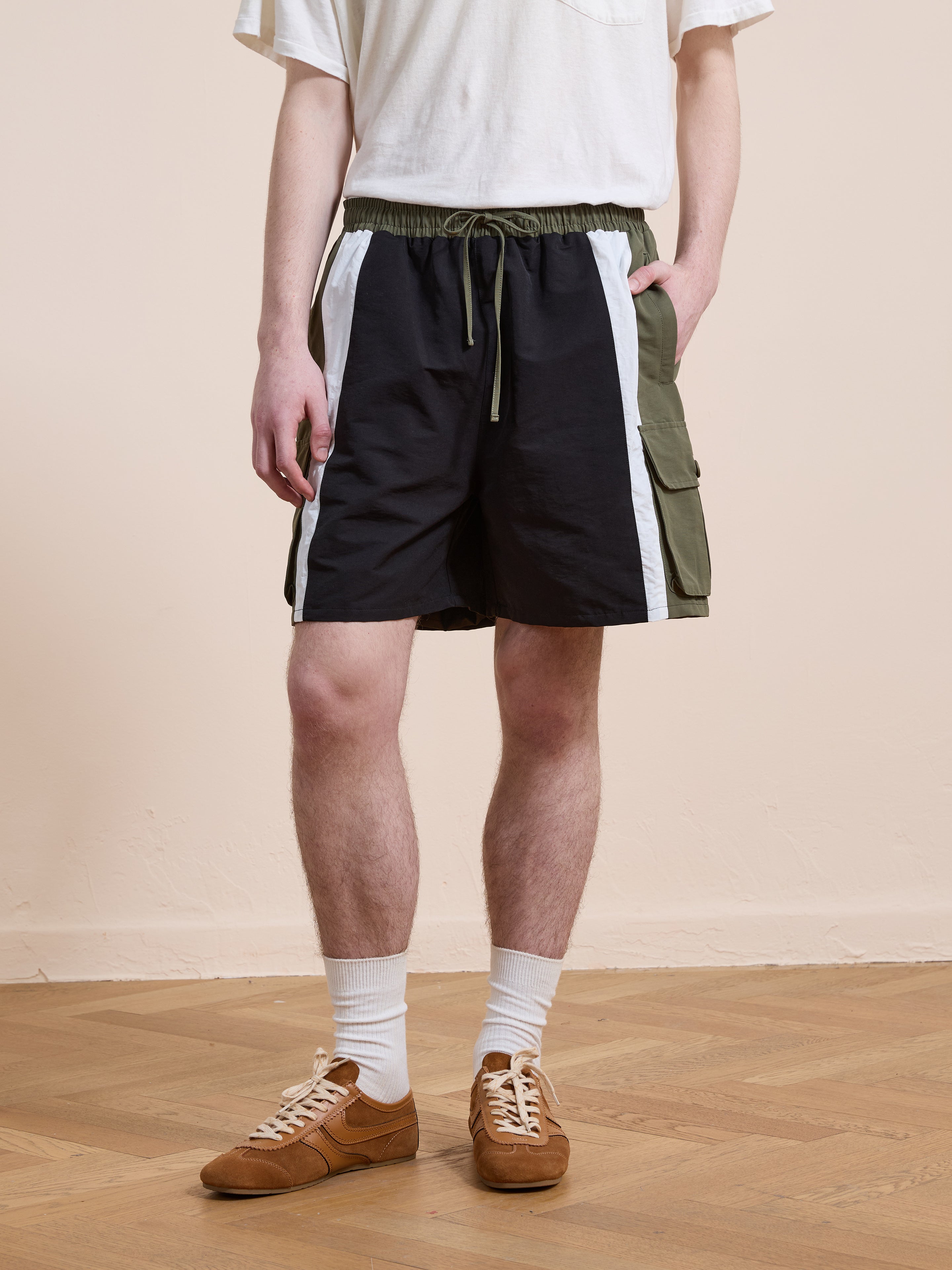 Someone wearing a white T-shirt and Found Sporting Club Track Shorts, featuring black, white, and green panels, along with white socks and brown sneakers, stands on a wooden floor.